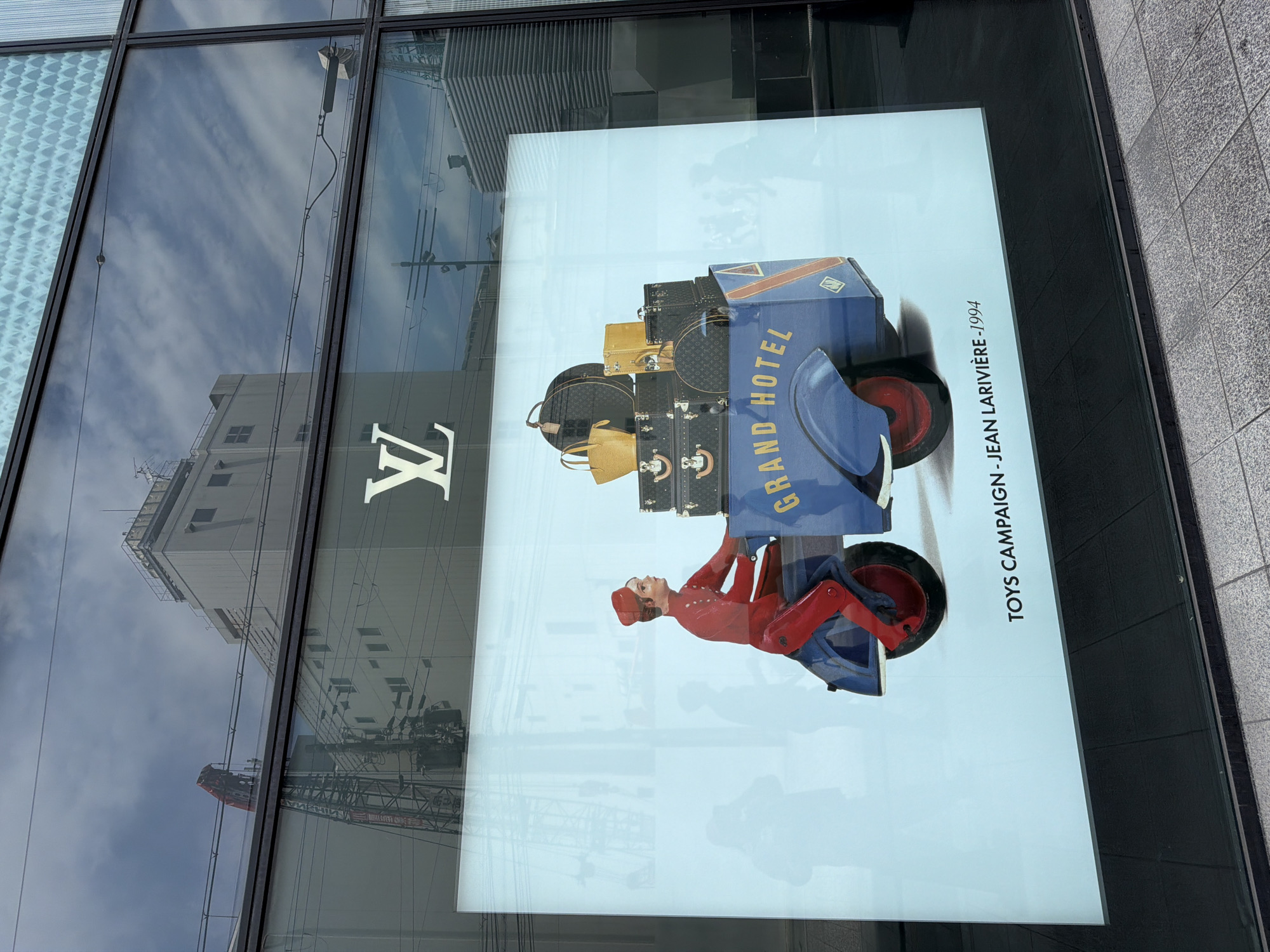 A large glass display window, likely for a Louis Vuitton store, is depicted at ground level in an urban setting, identified as Osaka, Japan, due to characteristic overhead utility lines and architecture. The window features a brightly illuminated advertisement behind the glass. The advertisement illustrates a stylized figure, a bellhop in a red uniform (red cap, jacket, and pants), riding a blue, three-wheeled vehicle resembling a scooter with a large cargo box. The box is inscribed with "GRAND HOTEL" in yellow script and is laden with several dark, monogrammed Louis Vuitton trunks and two yellow handbags. Below the illustration, black text reads "TOYS CAMPAIGN - JEAN LARIVIÈRE - 1994". A white Louis Vuitton "LV" logo is centered above the advertisement. The glass surface reflects a partly cloudy blue sky, a light-colored multi-story building with numerous windows, a prominent red-tipped construction crane, utility poles, and a dense network of overhead power lines. Faint, translucent silhouettes of pedestrians are visible in the reflections across the lower and middle sections of the advertisement. The ground immediately outside the window consists of a dark, flat surface transitioning to a lighter grey, textured sidewalk.