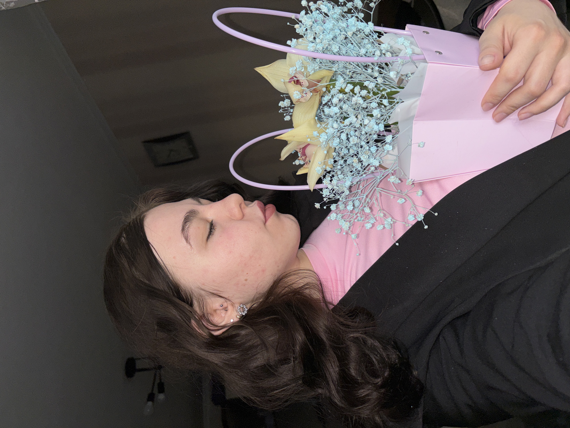 A woman with dark hair and closed eyes is lying down, holding a pink gift bag containing yellow orchids and blue baby's breath flowers. She is wearing a pink top and a black jacket. The location is Perm, Russia. A wall and a ceiling light fixture are visible in the background.