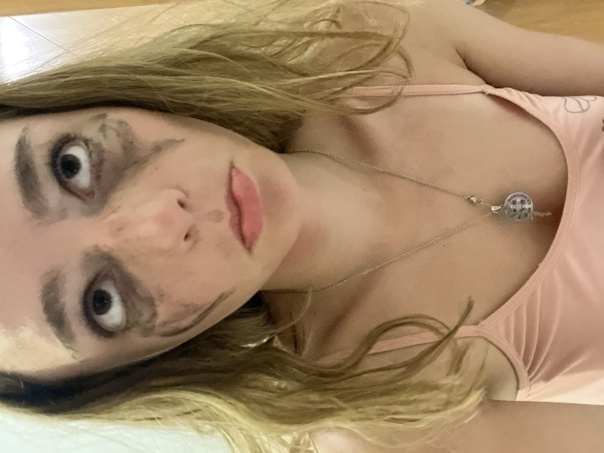 A close-up, angled selfie of a young woman with light blonde hair. Her face is prominently featured, with dark smudges around her eyes. She is wearing a light pink top with spaghetti straps and a silver necklace with a round pendant. The background is a light-colored surface, potentially a tiled floor. There is no information to confirm that the image was taken in San Bernardino Tlaxcalancingo, Mexico.