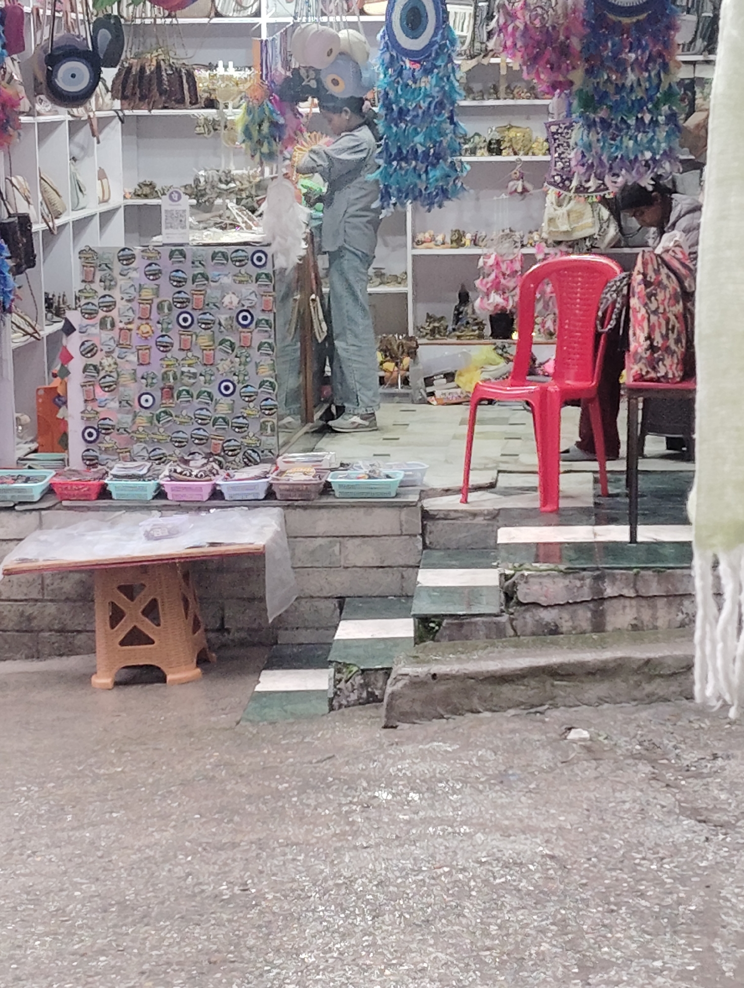 The image shows a market stall in Vashisht, India, with a variety of goods displayed for sale. In the background, shelves are stocked with items, some of which are obscured. In the center, a person in a gray jacket and blue pants is seen arranging merchandise on a stand. Several rows of small, colorful baskets are lined up in front of the stand, containing various items. In the foreground, a small, orange plastic stool sits next to a table covered with a white cloth. A bright red plastic chair is placed to the right of the stall, and a person is partially visible behind it. The ground appears to be a mix of stone and gravel. The overall scene depicts a bustling market environment with colorful decorations and numerous products.