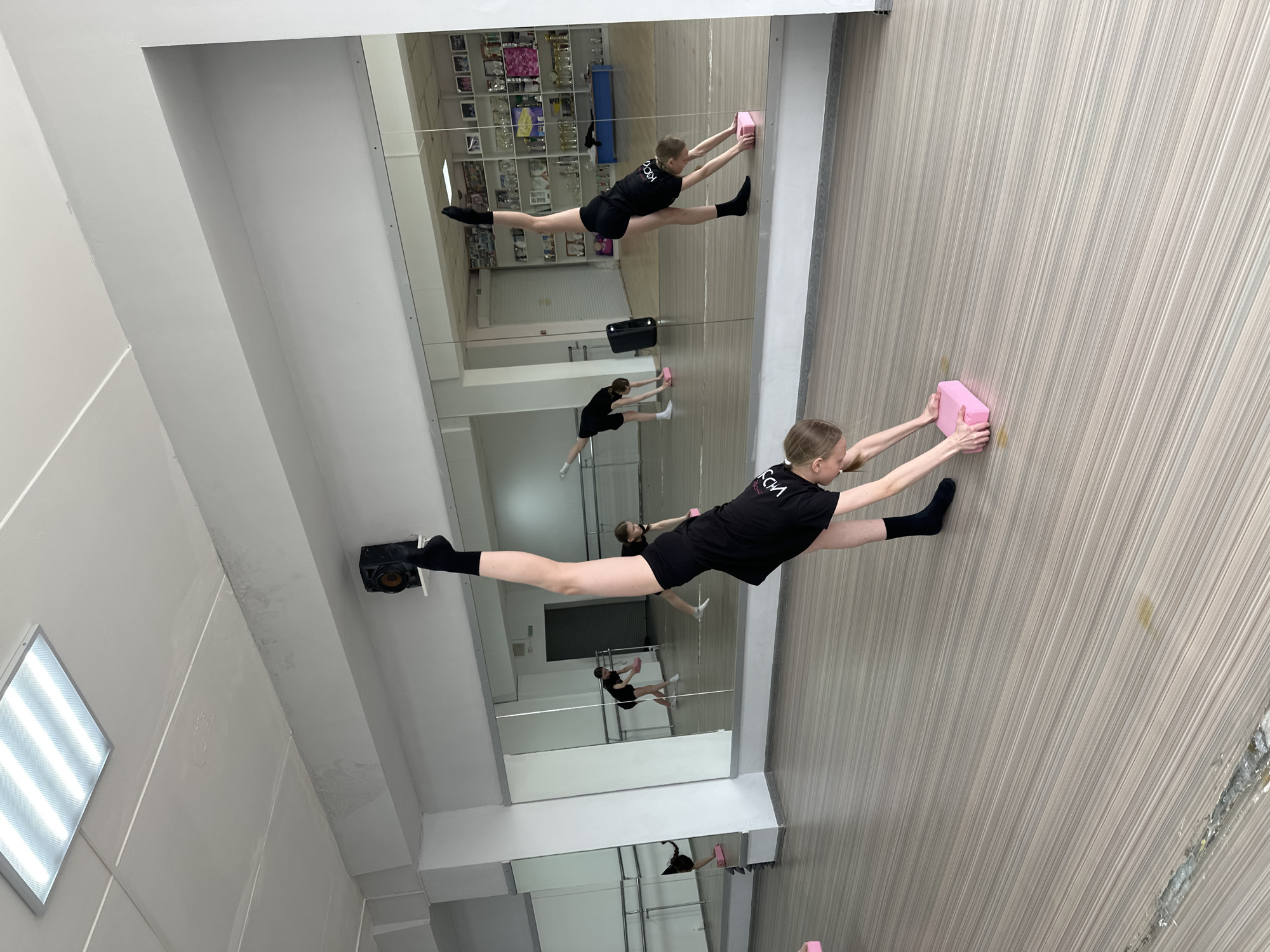 The image depicts a young female gymnast in a dance studio, as revealed by the mirrored walls, located in Votkinsk, Russia. She is in a straddle split position, with her left leg extended towards a mirrored wall and her right leg stretched towards a panelled wall. A pink foam block supports her hands on the right, while her left foot is braced against a light fixture. The studio has a white ceiling with recessed lighting. Other gymnasts are visible in the mirror, also practicing. The decor and equipment, including the foam block and mirrors, indicate a training environment.