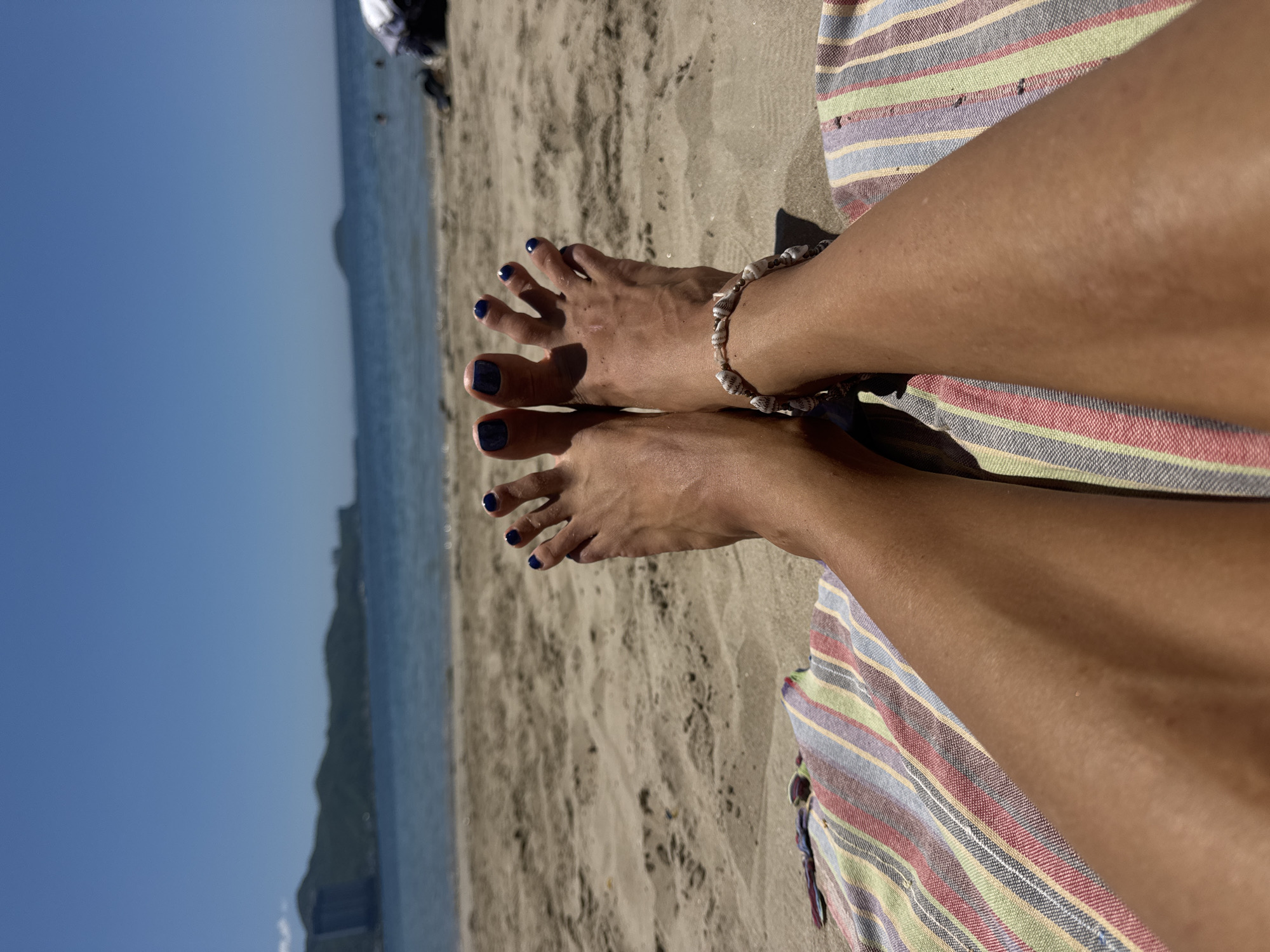A first-person perspective shot of a pair of tanned legs and feet, with blue nail polish on the toenails, resting on a striped beach towel on a sandy beach. The ocean and a distant, hazy coastline with mountains are visible in the background under a clear blue sky. The location is identified as Nha Trang Beach, Vietnam.