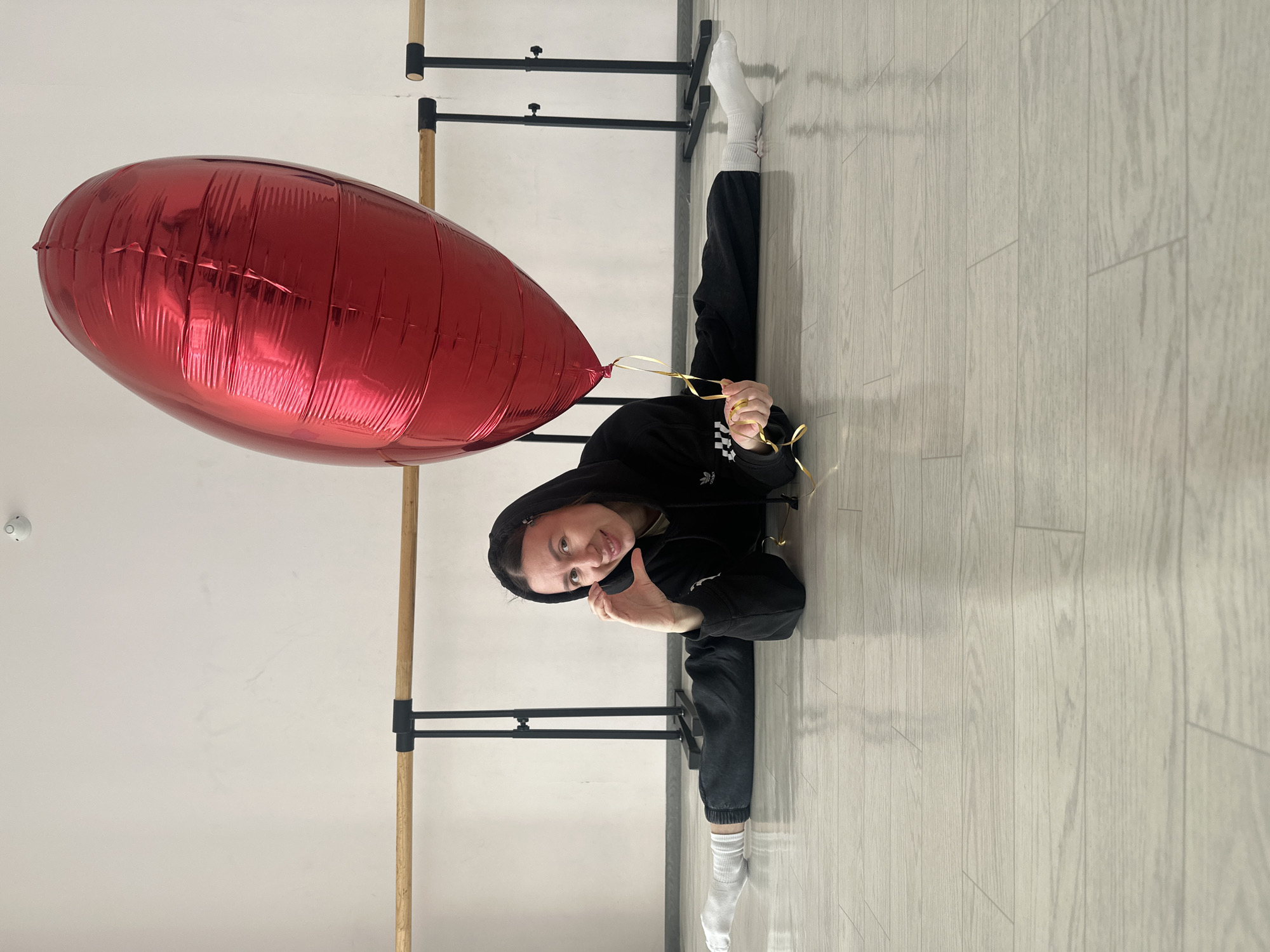 A woman in a black hooded sweatshirt and black pants performs a split in a dance studio in Votkinsk, Russia. She holds a large red heart-shaped balloon with gold ribbon. Her left leg extends towards a ballet barre fixed to the wall, while her right leg extends in the opposite direction, also towards the wall. She wears white socks on both feet. The floor is light-colored wood, and a small white security camera is visible on the wall. The scene is well-lit.