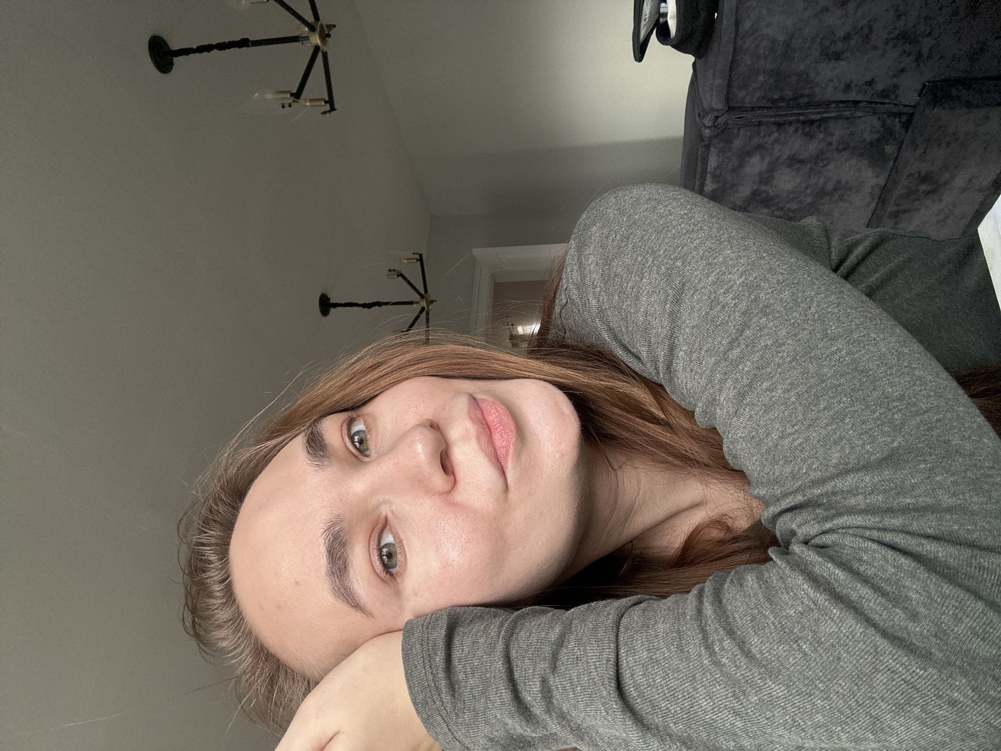 An indoor portrait captures a young woman with light brown or auburn hair and greenish-hazel eyes, looking directly at the camera with a relaxed and neutral expression. Her head is resting on her right arm, which is clad in a grey, long-sleeved, ribbed top. Her skin is fair, and her lips have a natural pink hue.
The setting is a room with light grey or off-white walls, likely a modern home interior in Kharnavy, Russia. Two black, contemporary chandeliers with multiple light sources are visible on the ceiling in the background. To the upper right, a section of a dark grey, textured sofa or armchair is partially visible, adding to the indoor residential context. A doorway further in the background suggests an adjacent room or hallway. The lighting is soft and ambient, characteristic of natural light from an unseen window, illuminating the woman's face gently. No text is visible in the image. The overall scene conveys a calm and casual moment.