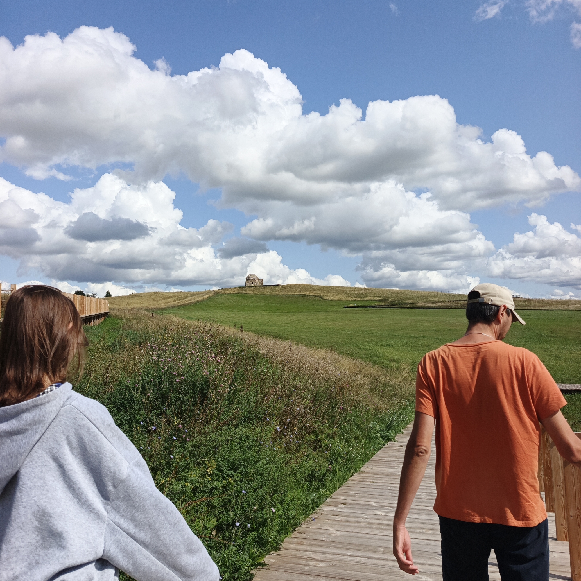 The image depicts two individuals walking on a wooden boardwalk in a rural, grassy landscape under a cloudy blue sky. The person on the left, seen from behind, has long brown hair and is wearing a gray hooded sweatshirt. The person on the right, also seen from behind, is wearing an orange t-shirt and a beige baseball cap.
In the distance, a small, stone, tower-like structure is visible atop a gently sloping hill. The surrounding environment consists of rolling green fields, tall grasses, and some wildflowers. The boardwalk appears to be elevated and curves to the right, suggesting a path through the natural terrain. The presence of the boardwalk and the structure on the hill indicate a possible park or historical site. This scene is located in the vicinity of Verkhnie Termy, Russia.