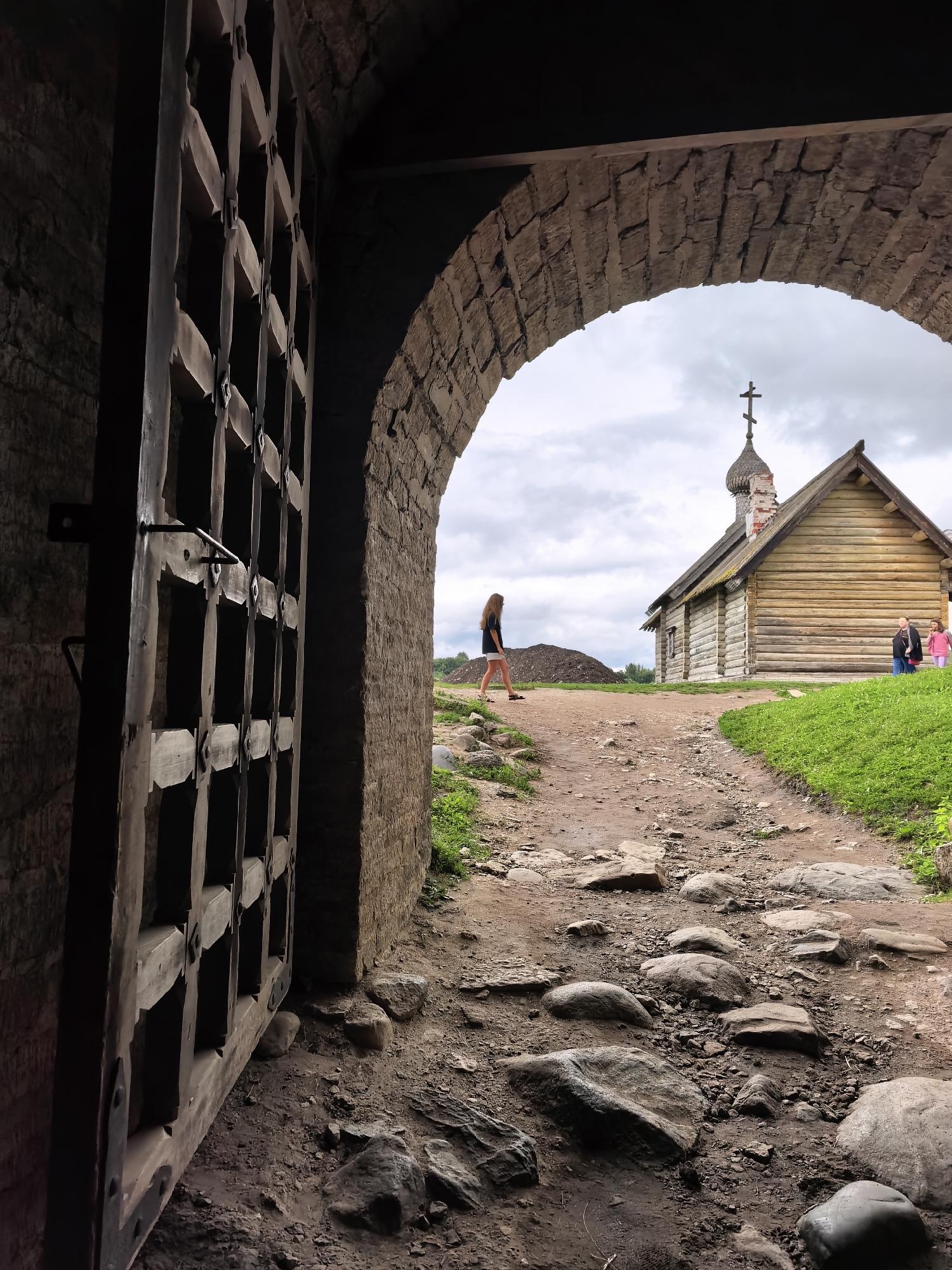 The image captures a view through a massive, weathered wooden gate and stone archway in Staraya Ladoga, Russia. The gate, constructed of thick wooden planks and metal fixtures, is partially open, revealing a rocky, dirt path leading towards a small, wooden church with a domed roof and a cross. The church is situated on a grassy knoll under a cloudy sky. A young woman with long, dark hair, wearing a black t-shirt and white shorts, walks away from the viewer on the path. Further down the path, near the church, two other figures, possibly children, are visible. The ground in the foreground is uneven, covered with large stones and loose dirt, suggesting an ancient or historical site. The stonework of the archway and the gate indicates a fortress or a similar historical structure.