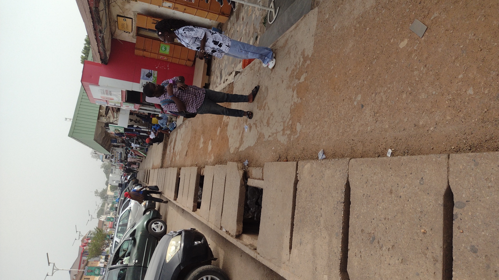 The image depicts a street scene in Jos, Nigeria. A gutter runs along the edge of the street, with concrete slabs providing intermittent coverage. Several cars are parked along the roadside, including a black SUV and a dark green car. A man in a plaid shirt and a woman in a patterned white shirt and jeans stand on the sidewalk near a building. In the background, shops line the street, and people are visible. The sky is overcast.