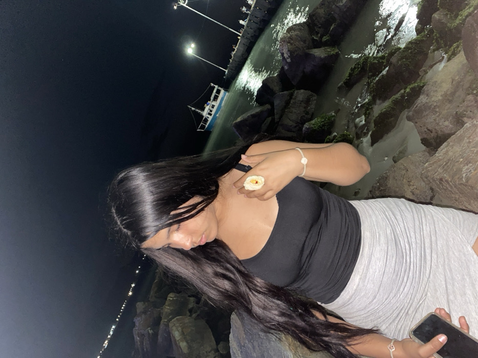 A woman poses on rocks at night near a body of water, likely the ocean. She wears a black tank top and grey skirt, holding a phone in one hand and adorned with a large floral ring. A dock and a boat are visible in the background, illuminated by streetlights. The lighting suggests it is dark, and the rocks near the water are coated in moss. The overall composition is angled and shot from a high perspective.