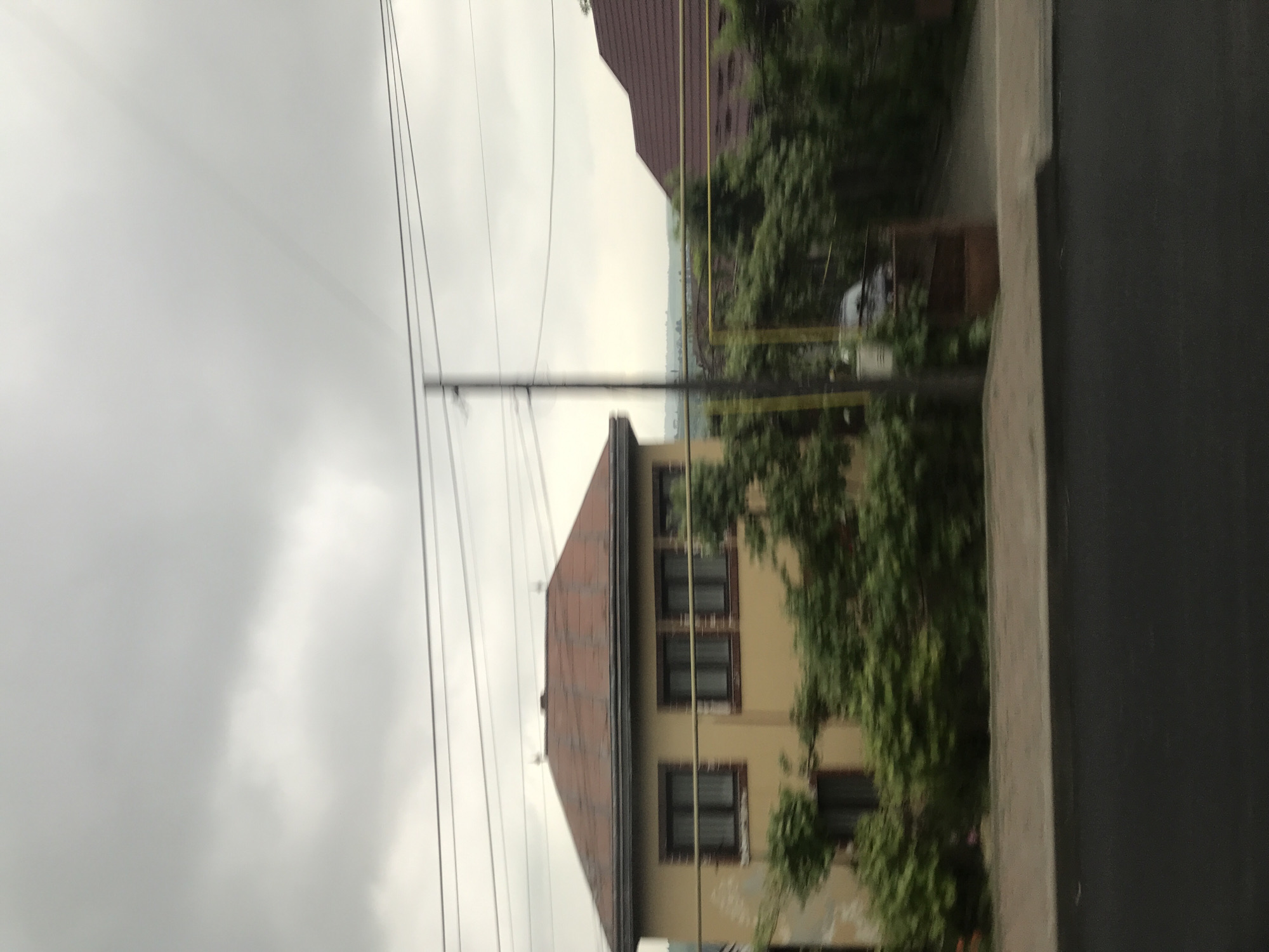 This image depicts a residential street scene in Fălticeni, Romania, under an overcast sky. The scene is observed from a slightly elevated, moving perspective, evidenced by motion blur, particularly in the foreground and peripheral elements.
The upper portion of the image is dominated by a light gray, cloudy sky, indicating overcast weather conditions. In the mid-ground, a hazy, light-colored horizon line is faintly visible between two houses, suggesting distant terrain or open landscape.
On the left, a two-story building with light yellow/ochre-colored stucco walls is prominent. It features a hip roof covered with reddish-brown, weathered tiles and dark trim along the eaves. Four rectangular windows with dark frames and light-colored curtains are visible on the upper level. A dark entrance or door is partially visible at ground level on the left side of the house.
To the right, partially obscured by a utility pole and vegetation, another building with a dark reddish-brown tiled roof is visible. Near this house, yellow horizontal utility pipes or structures are discernible behind dense green foliage.
A dark utility pole stands centrally, extending from the lower mid-ground towards the top. Multiple dark electrical or communication wires traverse horizontally across the upper sky, connected to the pole.
Dense green bushes and smaller trees are distributed across the mid-ground, situated in front of both houses and along the roadside. Some of this vegetation exhibits motion blur.
In the lower portion of the image, a dark asphalt or paved road surface is visible. Above the road, a light gray concrete sidewalk or curb runs horizontally. To the right of the utility pole, a dark, partially open gate, possibly made of metal or wood, leads into a property. A dark-colored car, likely a sedan, is partially visible behind this gate and surrounding foliage.
No individuals are visible within the frame. The overall environment suggests a residential area with established infrastructure and mature landscaping.