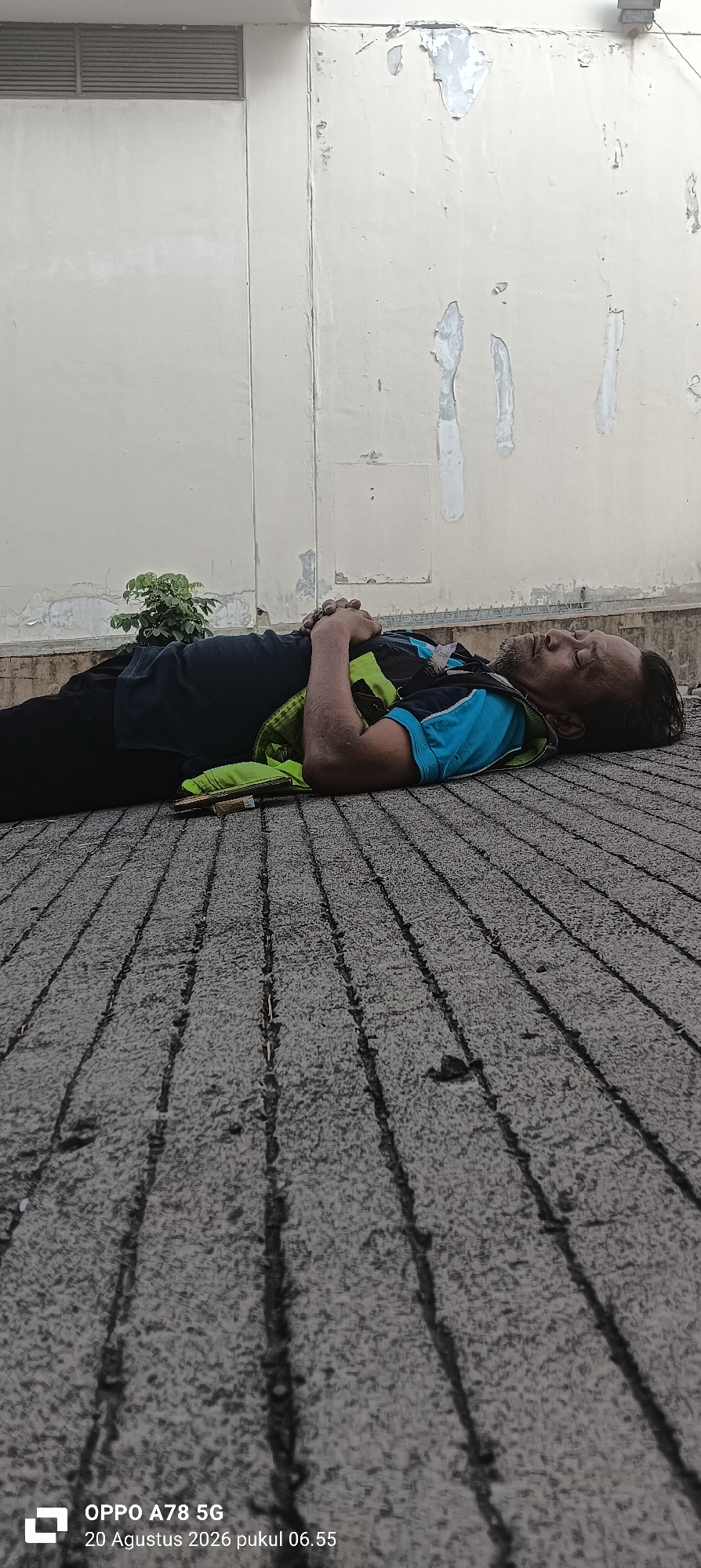 A man is lying on his back on a textured grey surface, possibly a paved outdoor area. He is wearing a dark blue t-shirt with a bright blue collar and a fluorescent green vest over it. His hands are clasped together over his chest. His eyes are closed and his head is tilted slightly back. There is a small green plant to his left. The background shows a light-colored wall with some peeling paint and a ventilation grate. The image is time-stamped "OPPO A78 5G 20 Augustus 2026 pukul 06.55" which translates to "OPPO A78 5G August 20, 2026 at 06:55". The location of Sedati Agung, Indonesia, is mentioned in the prompt, but not explicitly visible in the image.