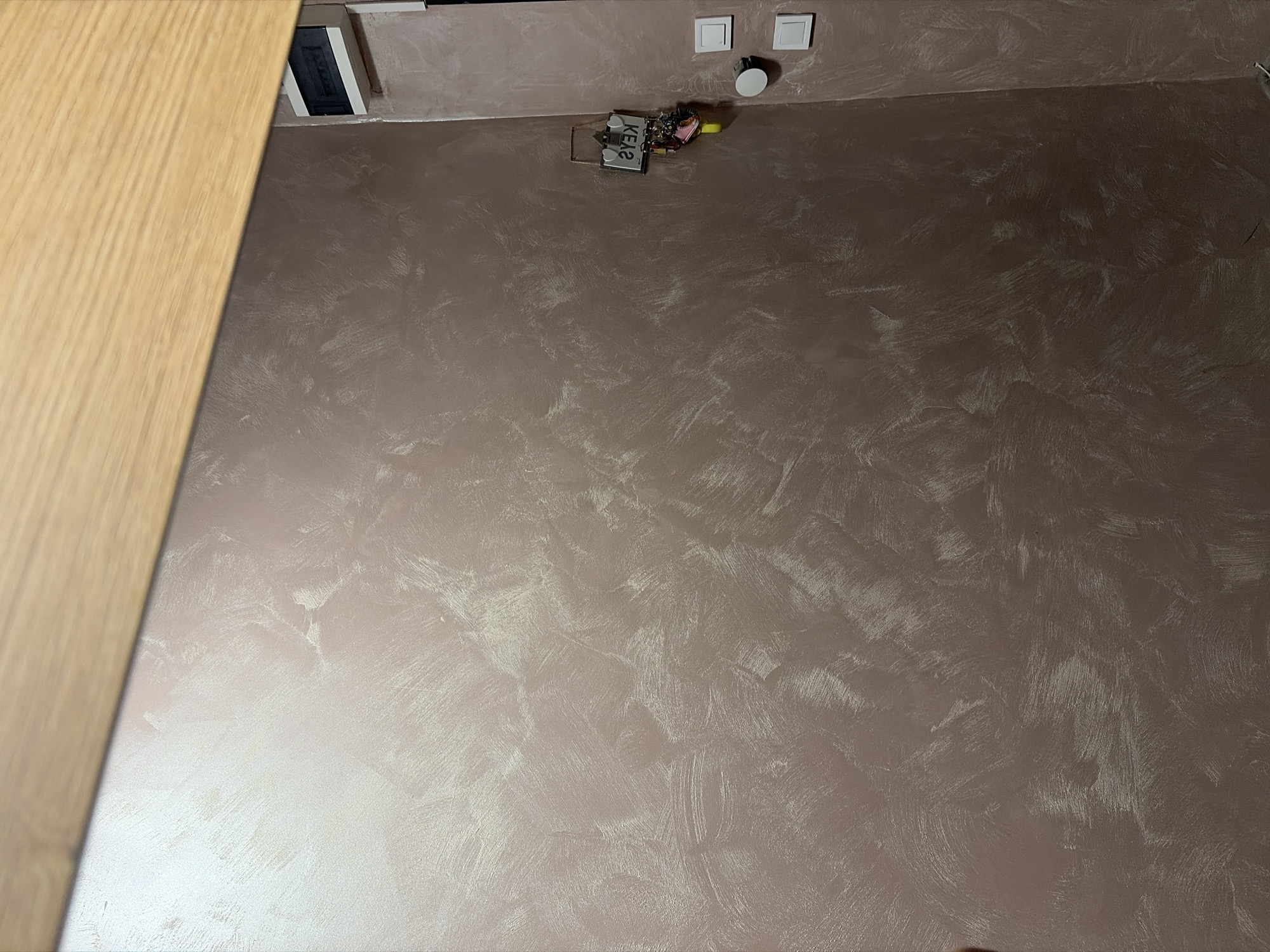 The image shows a section of an interior wall with a textured finish. The wall is a muted rose or taupe color with lighter, shimmering brushstrokes that create a marble-like or plaster effect. A wooden beam runs diagonally across the top of the frame, suggesting it might be part of a ceiling or shelf. On the right side of the wall, there is a key rack with several keys and a yellow charm hanging from it. Below the key rack are two light switches and a round electrical outlet cover. The lighting in the scene appears to be ambient, casting soft shadows and highlighting the texture of the wall. There are no people visible in the image. The environment appears to be residential or commercial, likely an indoor space such as a hallway or room.
