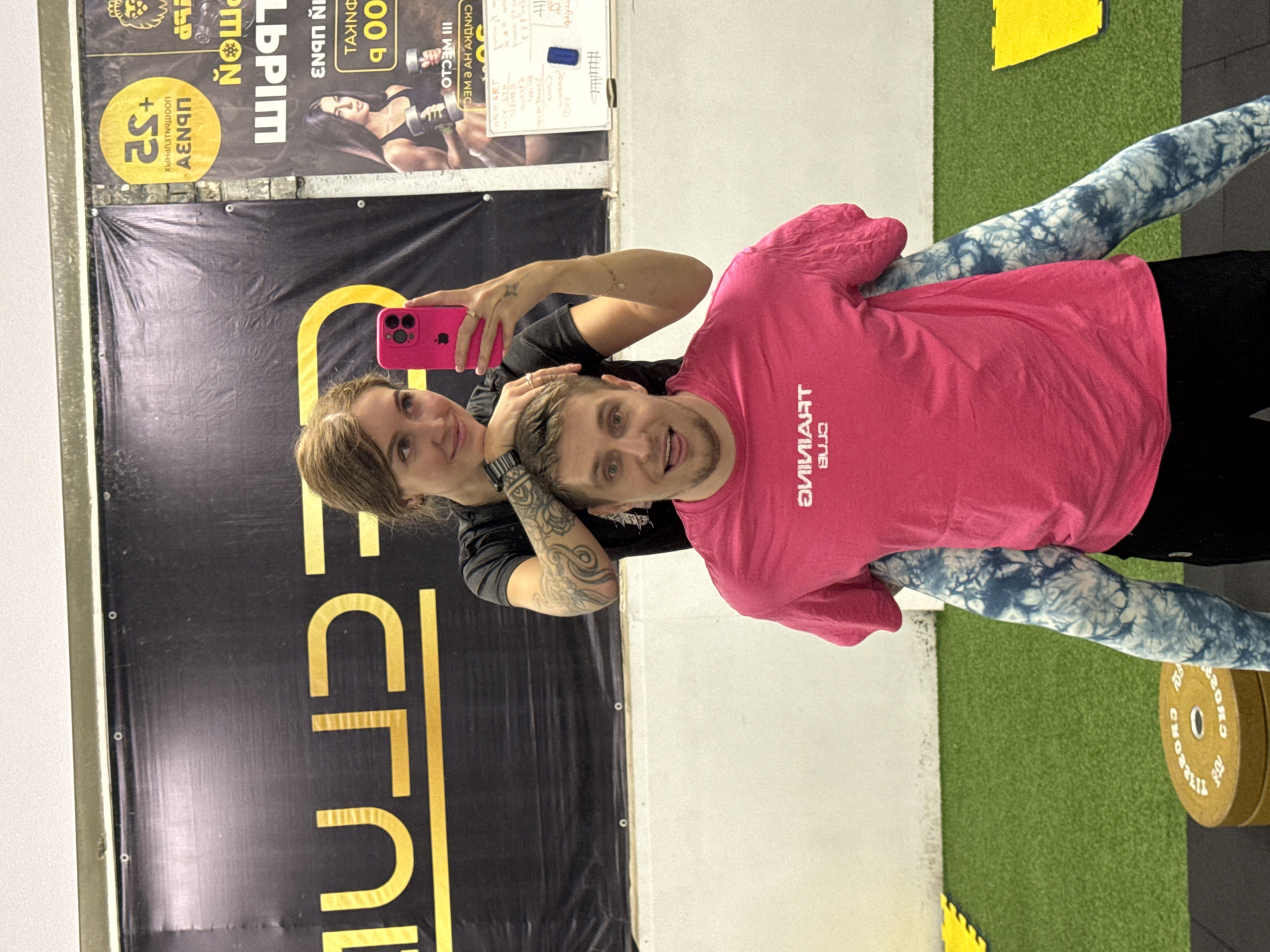 Here is a detailed description of the image:
**Scene Overview:**
The image captures two individuals, presumably a couple, taking a selfie inside what appears to be a gym or fitness center. The angle suggests that they are holding the phone together while taking the photo.
**People:**
1. **Woman:** Holding a pink phone, she is partially visible, with a focus on her face and the hand holding the phone. She has a tattoo on her arm.
2. **Man:** Wearing a pink shirt with mirrored text “TRAINING CLUB”, he is standing beside her. He has short hair and is smiling at the camera. He is wearing blue and white patterned leggings and black shorts.
**Subjects/Objects:**
* **Phone:** Pink-colored phone being used to capture the image.
* **Gym Environment:** Background includes gym equipment such as weight plates. Artificial green turf is visible. There's also a banner with promotional content.
* **Banner:** Shows an image of a woman lifting weights and some text, including the phrase “JOOMONIEUPHPIX +S2 АЕНОП” in reverse.
* **Weight Plates:** Yellow with white text.
**Actions/Interactions:**
* The primary action is taking a selfie.
* The man and woman are standing close together, suggesting an intimate relationship.
**Environment:**
The environment appears to be a gym or a training facility. There is a promotional banner on the wall and gym equipment is visible in the background.
**Location:**
The gym is located in Chaikovsky, Russia.