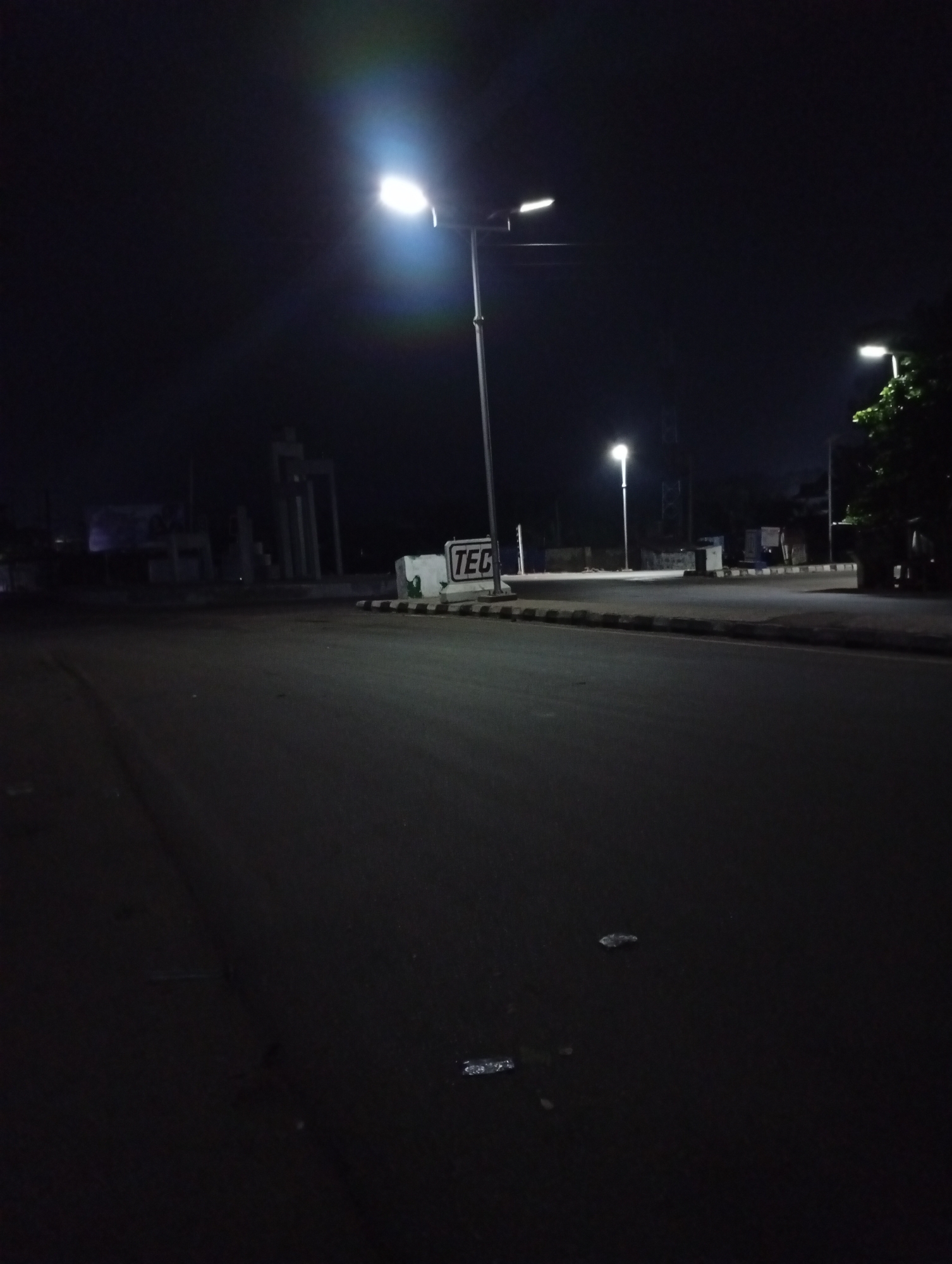 The image depicts an empty road at night in Adankolo, Nigeria. Streetlights illuminate the scene. In the background, there are buildings or structures, including a sign that reads "TEC". The road surface appears dark and possibly damp. There are no visible people or other subjects present. The overall environment appears urban and quiet.