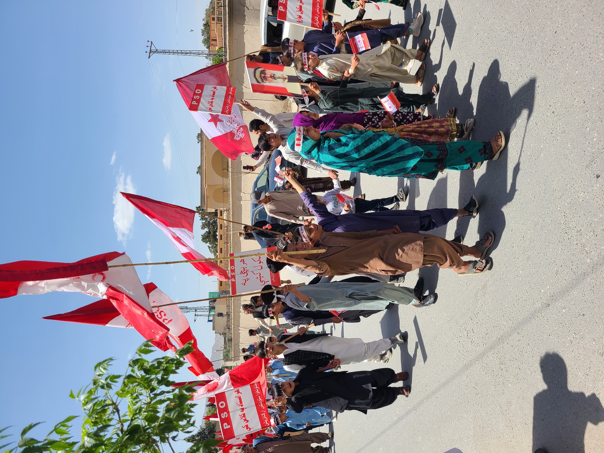 A group of people are marching on a paved road. They are holding red and white flags with the acronym "PSO" and Urdu text on them. Some people are also holding signs. The marchers are a mix of men and women, some wearing traditional clothing. The environment includes buildings and power lines in the background, and a tree in the foreground. The location appears to be Quetta City Tehsil, Pakistan. The sun is shining and casting shadows on the ground.