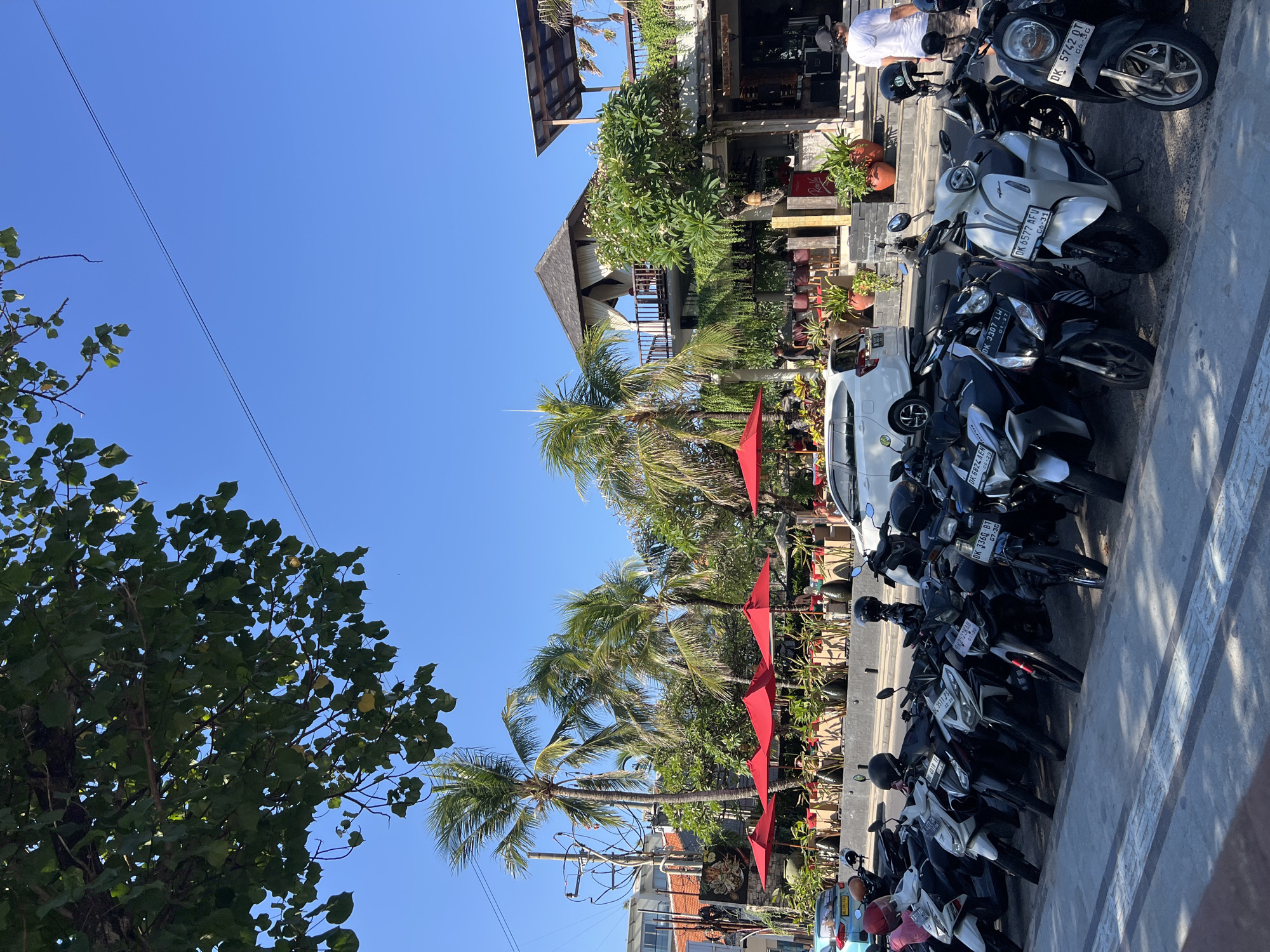 The image depicts a street scene in Kuta, Indonesia. In the foreground, a row of parked motorbikes lines a sidewalk. Beyond the motorbikes, a street is visible with several vehicles, including a white SUV. To the right of the street, a building with an outdoor seating area is visible, shaded by red umbrellas. Palm trees and lush green foliage surround the building. The sky is clear and blue. A person in a white hoodie and dark shorts walks away from the camera to the right.