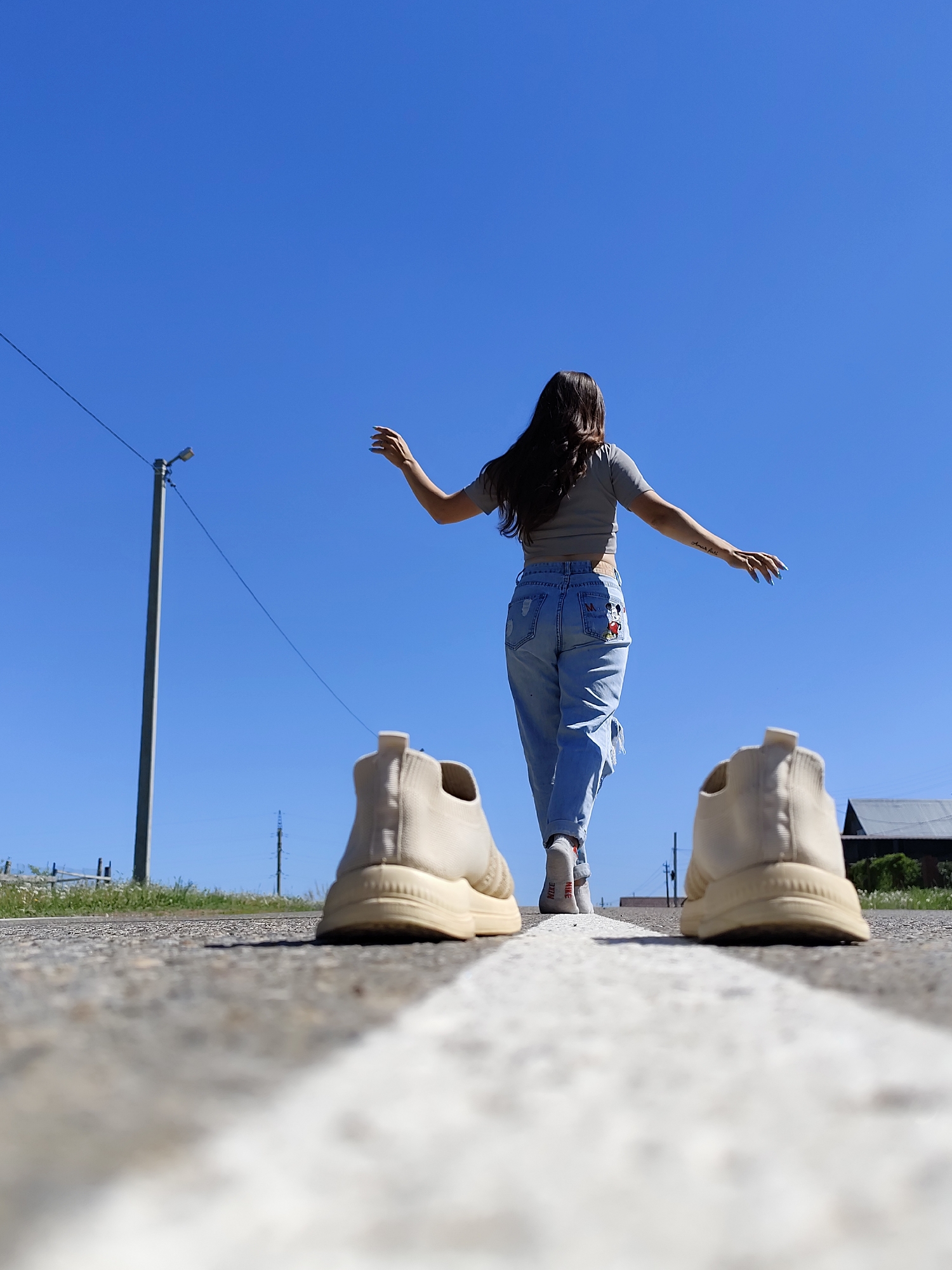 A young woman with long dark hair is walking away from the viewer on a road in Фоки, Russia. She has her arms outstretched and is wearing a grey short-sleeved t-shirt and distressed jeans with a Mickey Mouse design on the rear pocket. She is wearing white socks with the word "Nike" printed in red and has left a pair of tan sneakers on the white line in the middle of the road. The sky is a clear blue, and there is a street light on the left side of the frame. A building and some grass are visible in the background on the right. The road surface is grey asphalt.