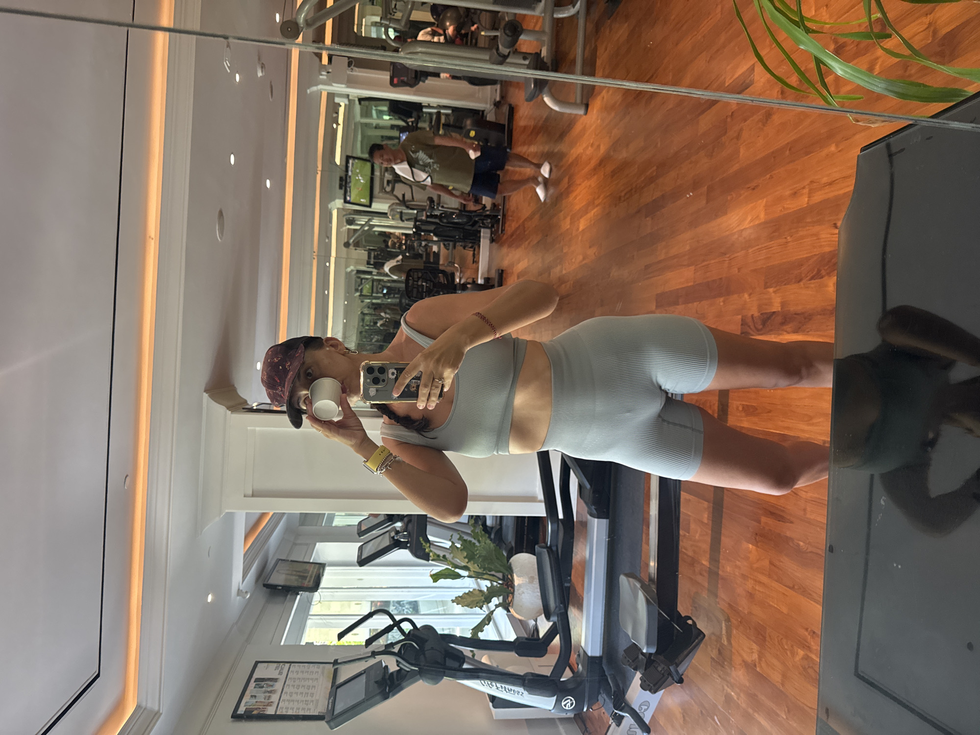 A woman is taking a mirror selfie in a gym in Kuta Selatan, Indonesia. She is wearing a light blue workout set and a maroon baseball cap. She is holding a white cup in one hand and a phone in the other. The gym has hardwood floors and various exercise machines. There are other people in the background working out. A plant is visible near the mirror. A reflection of the woman is visible in the mirror.