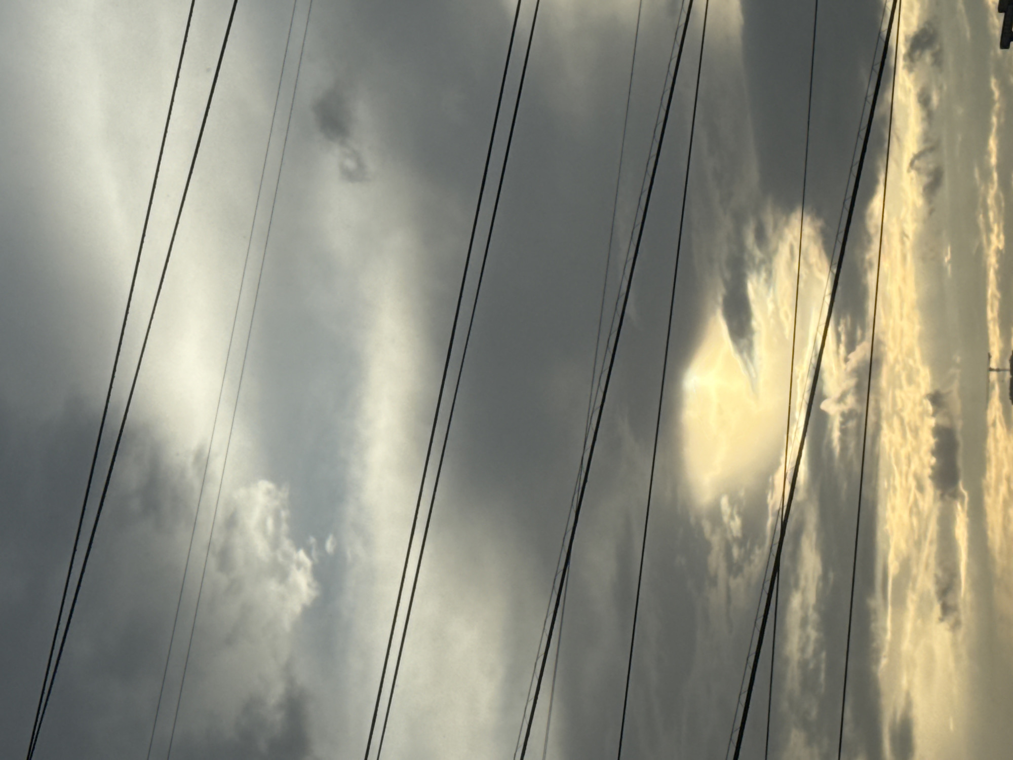 The image depicts a sky filled with clouds and several power lines stretching horizontally across the frame. The lighting suggests either dawn or dusk, with the sun partially visible through a break in the clouds, casting a warm, yellow glow. The clouds are varied in texture and color, ranging from dark gray to lighter, brighter white patches where the sun's rays are more direct. The power lines appear to be utility wires, common in urban environments. The lower portion of the image shows a sliver of the horizon, with the hint of structures that could be part of a cityscape. There are no people or distinct objects visible to definitively place this scene in Moscow, Russia, or any specific city. The overall impression is one of an overcast sky with a dramatic interplay of light and shadow, common during transitional times of day in many locations.
