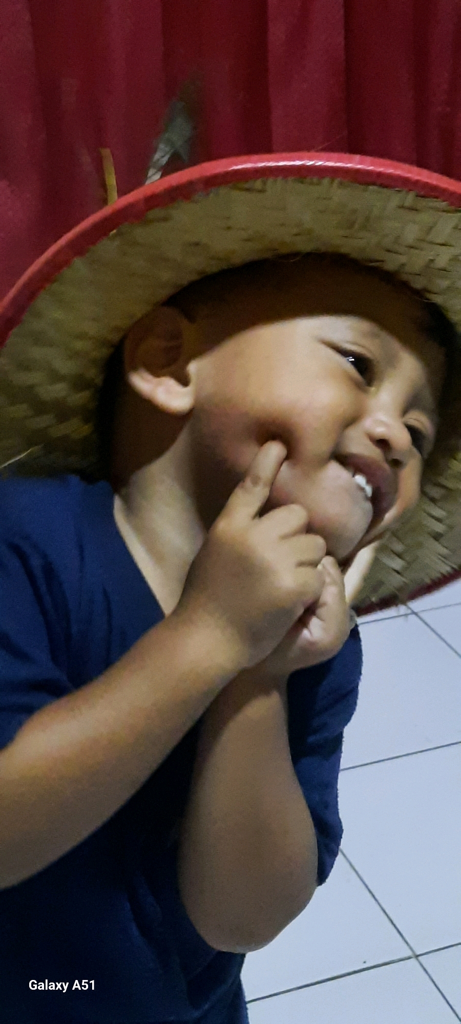The media file is a close-up, vertical shot of a young child, likely a boy, wearing a straw hat with a red rim. The child is wearing a dark blue shirt. The child's hands are positioned near their face, with one finger gently touching their cheek, possibly to emphasize their dimples. The child's face is turned to the side, with a bright smile showing teeth and eyes that are looking slightly upwards. The background consists of a red curtain and a tiled floor. The lighting suggests an indoor setting.
Notable details include the child's joyful expression and the way they are interacting with their own face. The straw hat is a prominent object. The setting appears to be a home, given the domestic background elements. There is no indication of the time of day or weather.
Visible text includes "Galaxy A51" in the bottom left corner, indicating the device used to capture the image. The location context is Ploso, Indonesia.