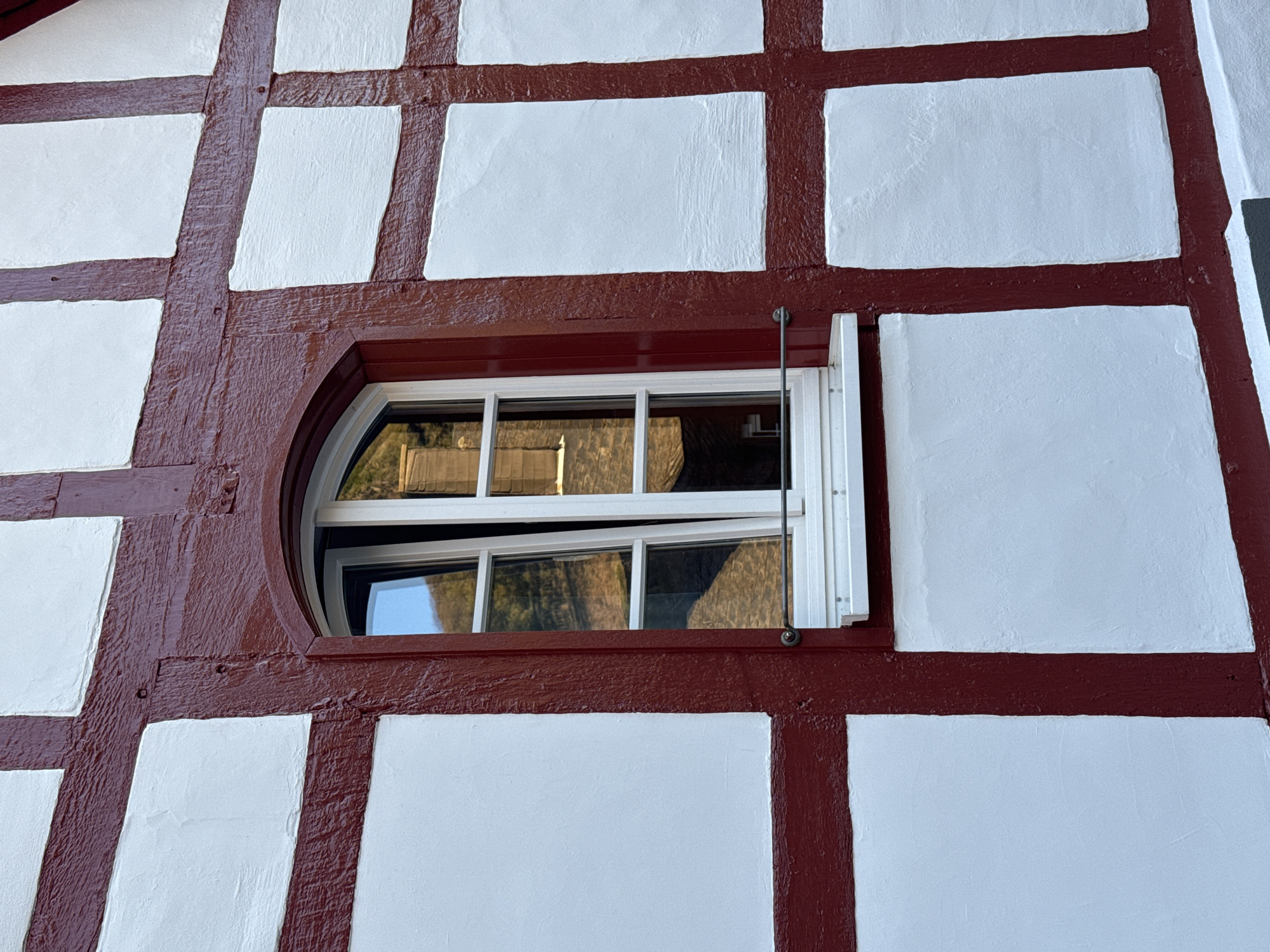 A low-angle, medium shot depicts a section of a building with a half-timbered (Fachwerkhaus) design, typical of architecture in Beilstein, Germany. The structure features a grid of dark red, roughly hewn timbers filled with white plaster panels. In the center, a horizontally oriented oval-shaped window sits within the timber framework. The window's white frame holds multiple panes and is slightly ajar, revealing a glimpse of the exterior reflected in the glass. A thin metal rod extends from the upper frame. The timbers show signs of weathering and age. The overall scene evokes a sense of traditional European architecture and craftsmanship.