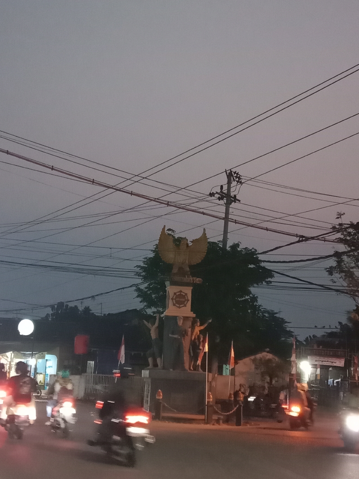 The image depicts a monument in the city of Langensari, Indonesia, at dusk. The monument features a golden eagle statue at the top, with figures of people at its base. Several Indonesian flags are displayed near the monument. The foreground shows a street with several motorcycles in motion, their headlights and taillights blurred due to movement. People are visible riding the motorcycles. In the background, there are trees, buildings, and power lines crisscrossing the sky. The overall atmosphere suggests a busy street scene in an Indonesian city during twilight.