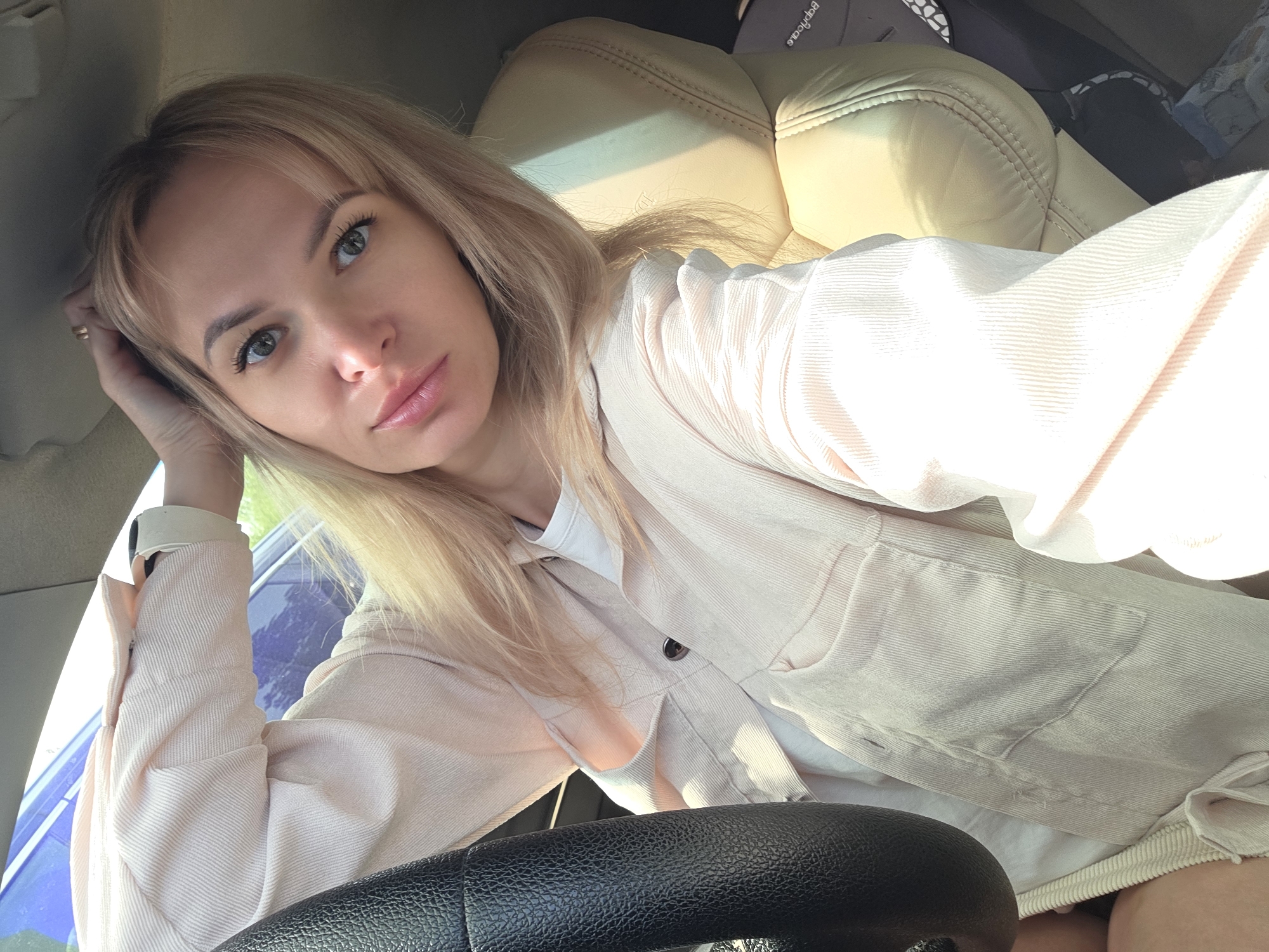 The image depicts a woman in the driver's seat of a car. She has blonde hair, light skin, and is wearing a light-colored corduroy jacket over a white shirt. Her right hand is resting on her head. The steering wheel is visible in the foreground. The car seat is light beige. The image appears to have been taken inside a car, possibly in the city of Chaikovsky, Russia.
