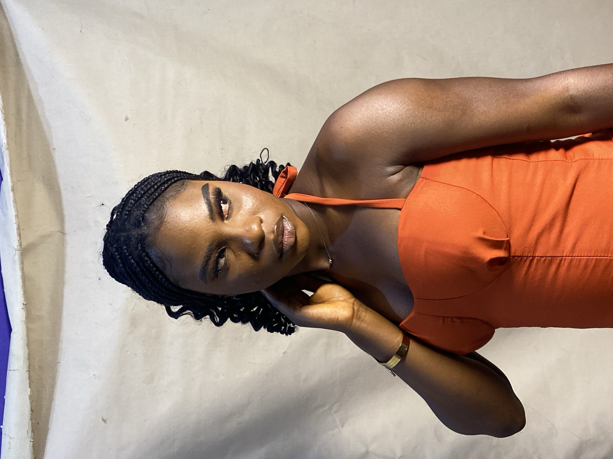 A medium shot, portrait-style, presents a dark-skinned woman wearing an orange top. Her hair is styled in braids with curled ends. She wears a gold bracelet on her left wrist and a gold necklace. Her left hand is raised to her face, resting on her jawline. The background is a light, neutral color. Without further context, it's impossible to determine the precise location in Jos, Nigeria.