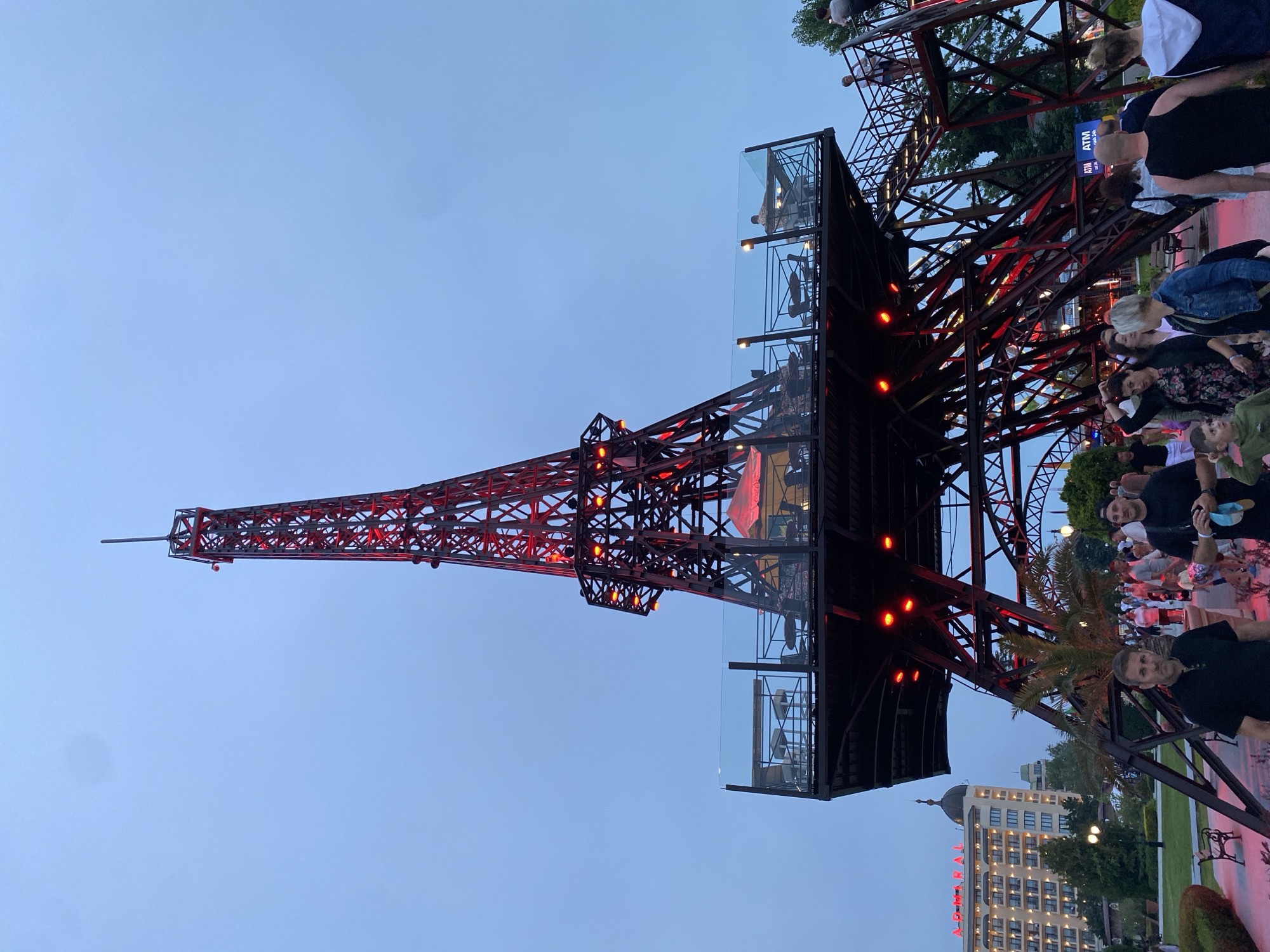 The image captures an evening scene in Golden Sands, Varna, Bulgaria, featuring a large, illuminated replica of the Eiffel Tower. The sky is clear with a light twilight blue hue.
The central subject is a dark metal lattice structure resembling the Eiffel Tower, internally lit with red lights. Midway up the structure, an expansive open-air platform serves as a restaurant or viewing deck, enclosed by glass railings. Tables and chairs are arranged on this platform, with several patrons visible. Red umbrella-like structures are present on the platform. Stairs ascend to this elevated area from the ground level on the right.
At the base of the tower and extending into the mid-ground, a paved pedestrian area is populated by numerous individuals of varying ages, casually dressed, walking and standing. Green spaces with manicured hedges, bushes, and trees, including a prominent palm tree, surround the walkway. Near the base of the tower on the right, two blue "ATM" signs are visible.
In the background, a large, multi-story hotel building with a light-colored facade and numerous windows is prominent. The top of this building features a distinctive domed structure. The name "ADMIRAL" is displayed in red, illuminated letters on the building's facade. The overall lighting suggests dusk or early night, with artificial lights illuminating the tower, the hotel sign, and some ground areas.