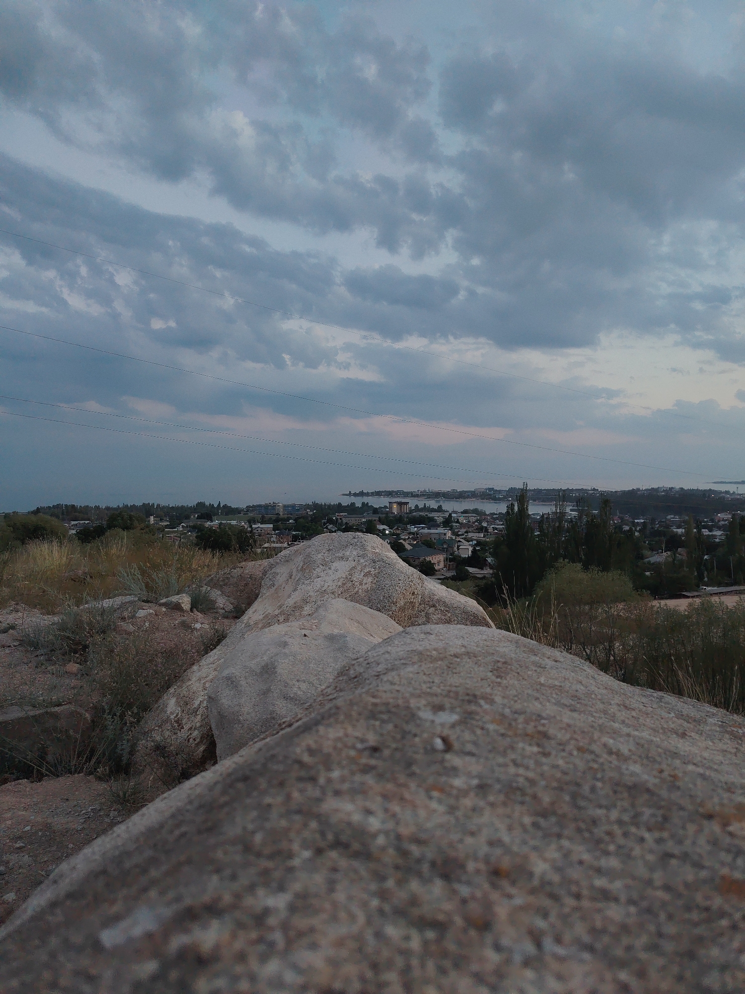 This image captures a panoramic view of Cholpon-Ata, Kyrgyzstan, from a rocky vantage point. In the foreground, large, weathered boulders dominate the composition, covered in sparse, dry vegetation. Beyond the rocks, a town sprawls towards a body of water, likely Issyk-Kul Lake, which is visible in the distance as a hazy blue expanse. The sky above is overcast with dramatic, layered clouds in shades of grey and blue, with thin power lines crisscrossing the upper portion of the frame. The overall scene suggests an early evening or late afternoon setting, with soft, diffused light. The town consists of a mix of low-rise buildings and scattered trees, with some taller structures and more dense foliage visible closer to the water's edge. There are no people or visible actions in the scene.