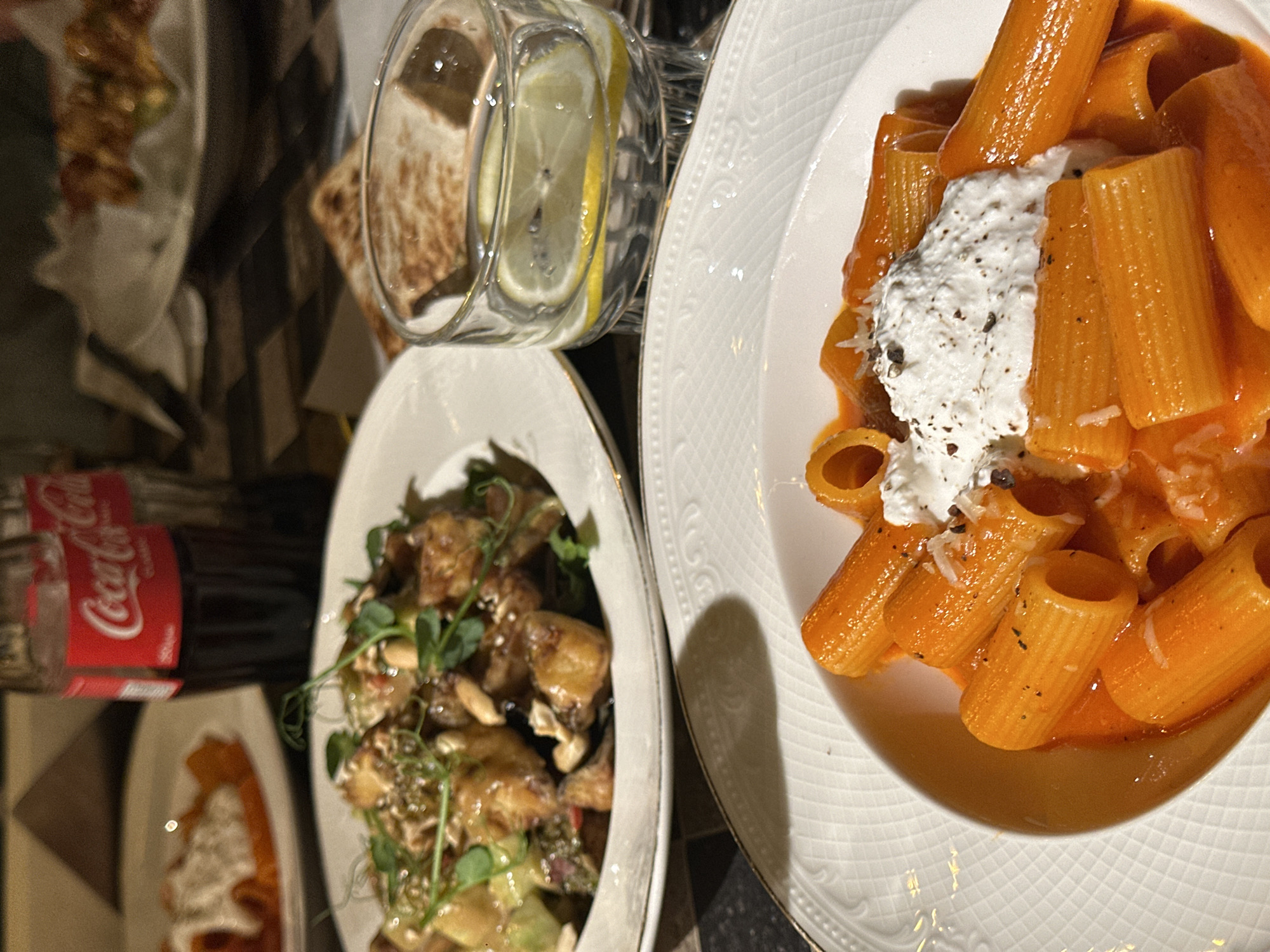 The image shows a table setting with various dishes. In the foreground, there is a white plate with rigatoni pasta in a tomato-based sauce, topped with a dollop of ricotta cheese and some spices. Next to it is a white bowl containing a dish with what appears to be eggplant, nuts, and sprouts.
Behind the pasta plate and bowl, there is a glass of water with a lemon slice and a bottle of Coca-Cola. There are also other dishes partially visible on the table. The table appears to be set in a restaurant.
Based on the dishes and the style of the setting, the restaurant could be located in Yerevan, Armenia, known for its diverse culinary scene, as requested. However, there is no explicit visual evidence to confirm this location.