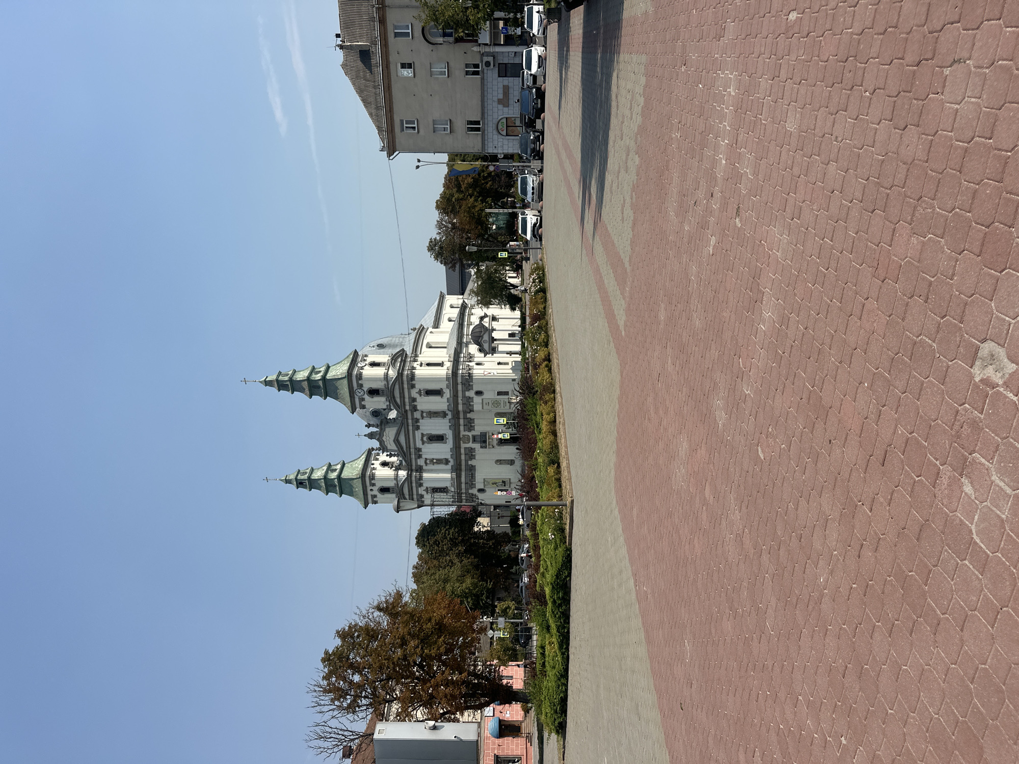 The image depicts a wide, paved square in Ternopil, Ukraine, dominated by a large, ornate white church with two distinctive green spires. The square is paved with red and grey interlocking bricks, leading the eye towards the church. Several cars are parked along the street in front of the church and adjacent buildings, which include a multi-story apartment building and a smaller pink building. Trees with autumnal foliage are scattered throughout the scene, and a neatly landscaped garden area with bushes and flowers is situated in front of the church. The sky is clear and blue. The scene is tranquil, with no people prominently visible in the frame, suggesting a quiet moment in the city.