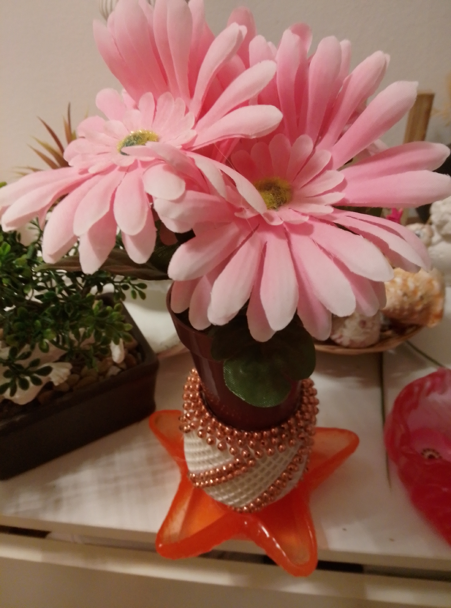 A close-up, eye-level shot shows a bouquet of artificial pink gerbera daisies in a brown ceramic pot that sits on an orange star-shaped base. The flowers are the primary focus, with their delicate pink petals and bright yellow-green centers. To the left of the pot, a dark brown square planter contains green artificial foliage and small white shells. The background is a soft, neutral tone, suggesting an indoor setting. The image is well-lit, highlighting the textures and colors of the floral arrangement.