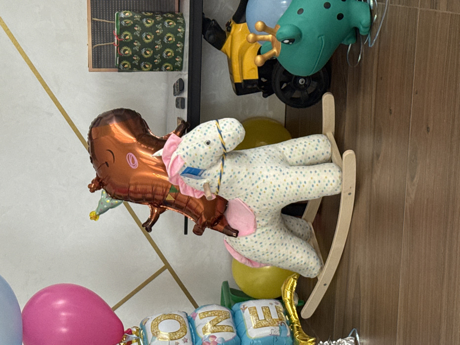 An indoor scene, possibly a room decorated for a child's celebration, is depicted in Sibir', Russia. The foreground features a plush rocking horse with a light-colored, patterned fabric body, pink mane, and pink saddle, resting on a light wooden rocker base. A blue and yellow braided string is around its snout, and a small white tag hangs from its neck. To the left, a cluster of balloons includes three light blue square balloons spelling "ONE" vertically with gold outlines and stars, topped by a solid pink round balloon, and a partial light blue balloon at the upper left. A silver foil balloon weight is at the base of the "ONE" balloons.
Behind the rocking horse, a large, shiny, copper-brown foil balloon shaped like a dinosaur, wearing a small green and yellow party hat, stands upright. This balloon features closed eyes and a smiling mouth. Several yellow round balloons are partially visible behind and around the rocking horse and dinosaur balloon.
On the right side of the image, partially obscured by the rocking horse, a dark brown wooden floor is visible. Further right on the floor, a green toy frog-like figure with black spots and a gold crown is visible, resting on clear plastic runners. Behind it, a yellow and black toy vehicle with black wheels is partially visible.
In the background, a light-colored wall features thin, intersecting gold diagonal lines. A dark shelf against this wall holds a rectangular object resembling a black pegboard with a brown frame, a green gift bag with a repeating white wreath pattern and red accents, and two small dark objects resembling remote controls. A light blue round balloon is partially visible near the green toy and the shelf. The scene is static, with no human figures or active interactions present.
