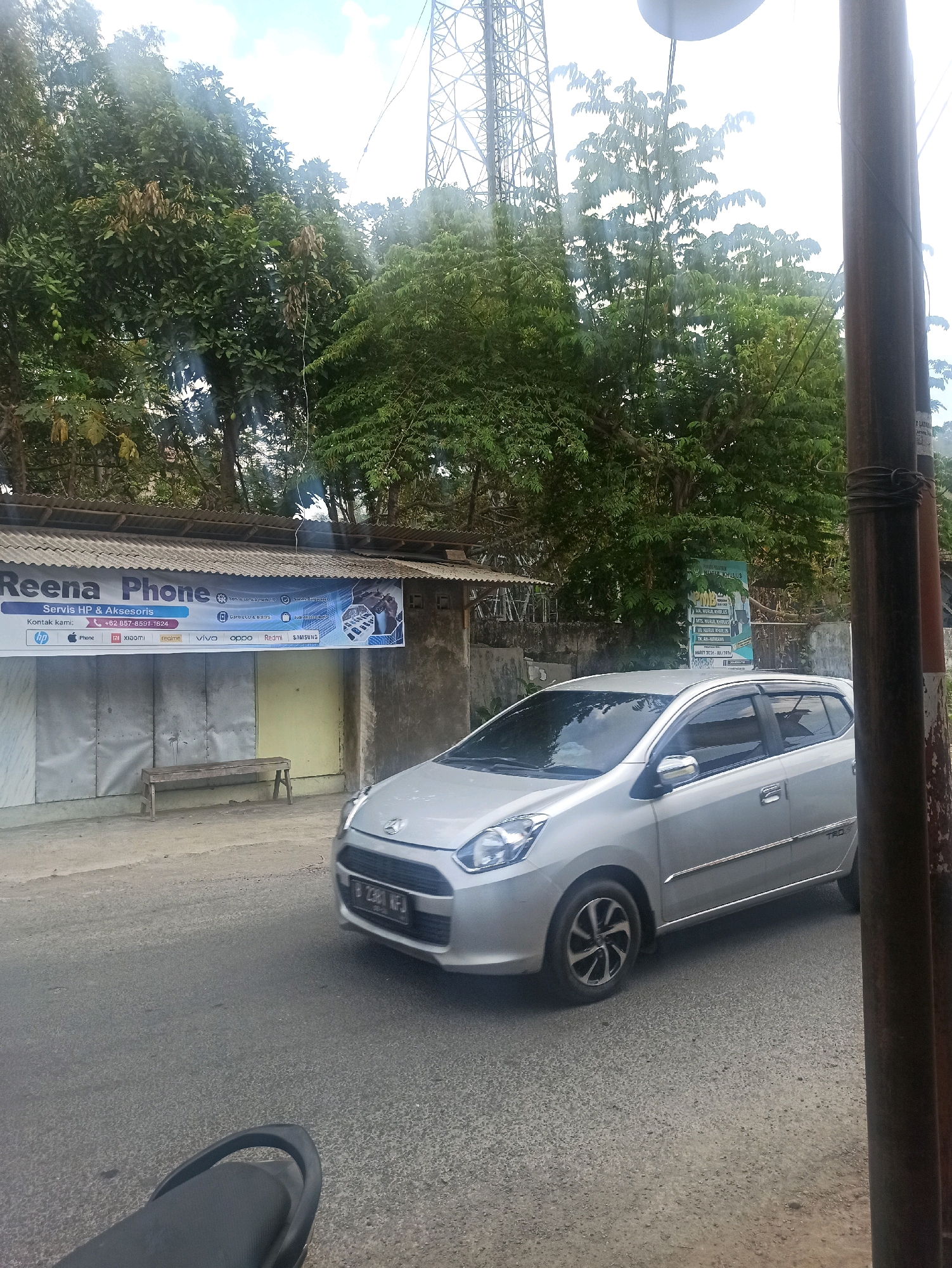 A silver car drives on a paved road in front of a row of shops. The car is a hatchback with a license plate that reads "B 2381 KFJ". One shop has a sign that says "Reena Phone". Behind the shops, there are trees and a tall tower, possibly a cell tower. The sky is partly cloudy. There is also a motorcycle seat in the foreground on the left side of the image. The overall scene appears to be a street in Tlogosari, Indonesia.