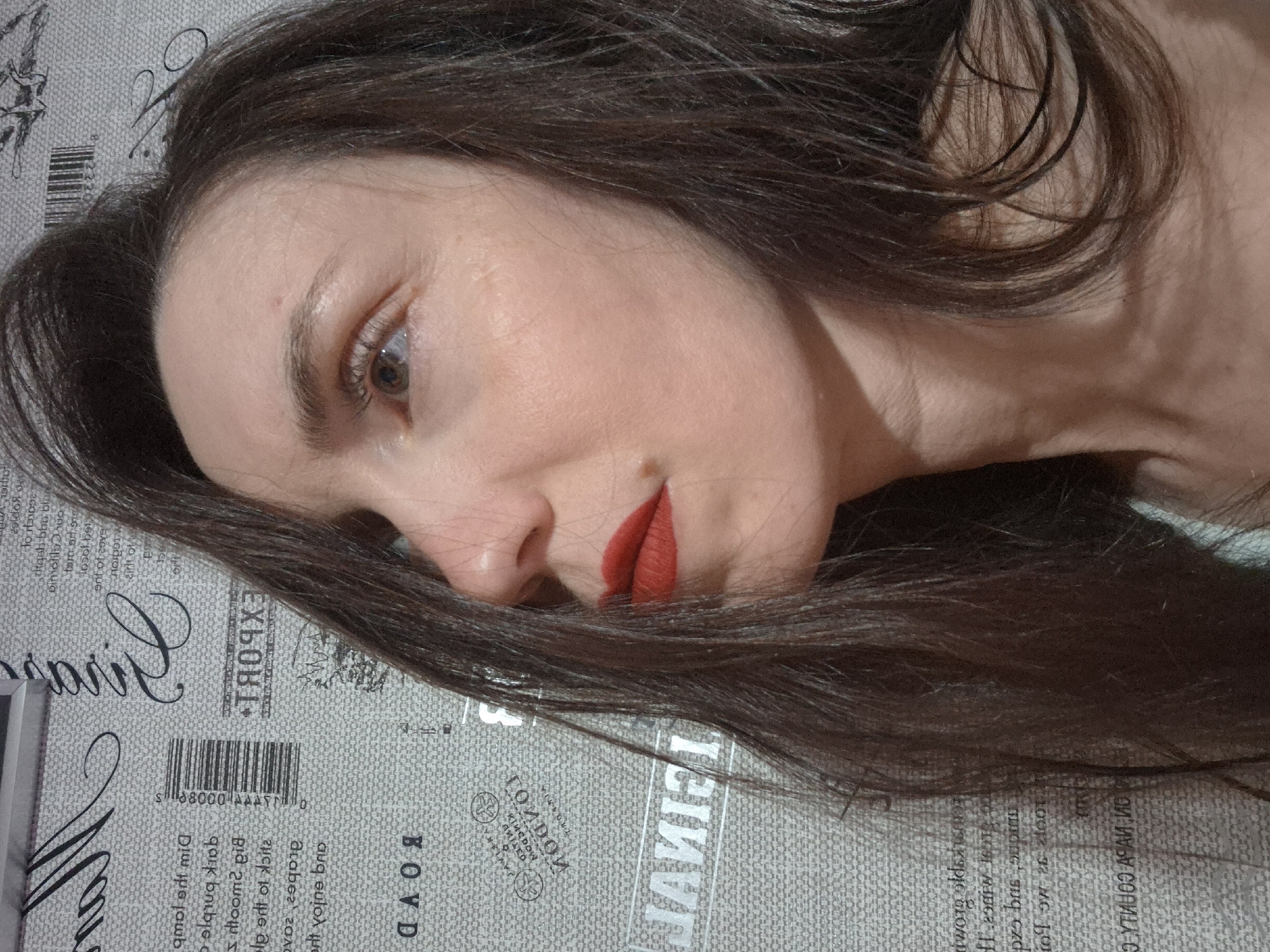 The image shows a woman with brown hair lying on a surface with printed text, possibly a sheet or cover. Her head is tilted to the side, and she is wearing red lipstick. The text on the surface includes words like "EXPORT," "LONDON," "FINANCIAL," and "BOYD." A barcode is also visible. The setting and specific location are not identifiable from the image.