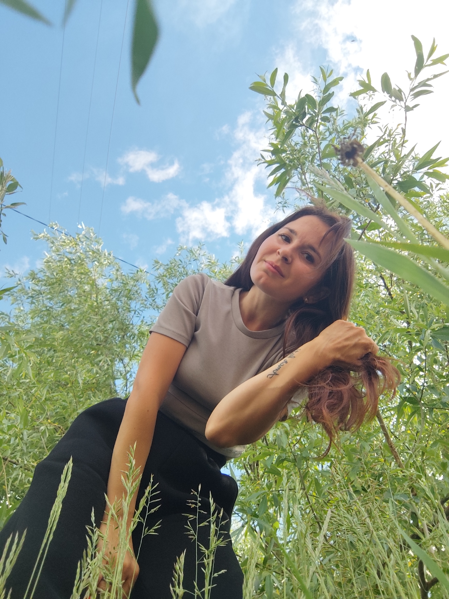 A low-angle outdoor shot captures a woman amidst lush green foliage and a bright blue sky with scattered white clouds. The camera is positioned on the ground, looking upwards. The woman, with long, reddish-brown hair, is wearing a light taupe t-shirt and what appears to be a black skirt or loose pants. She is leaning into the frame, her head slightly tilted, and she looks directly at the camera with a calm, subtle expression. Her right arm is visible, with a cursive text tattoo on her inner forearm that reads "in comfort". She is gently holding a lock of her hair with her right hand.
The background is dominated by a dense canopy of green leaves, possibly from willow trees given their slender shape, and tall green grasses that extend into the foreground, framing the shot. Several thin power lines are visible crossing the upper left portion of the sky. A prominent dried seed head or plant stalk is seen in the upper right, close to the camera, adding depth to the foreground. The scene is brightly lit by natural daylight, suggesting it is daytime. The overall setting evokes a natural, serene environment in Фоки, Russia.