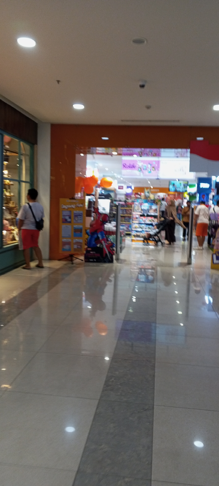 The image captures a wide-angle view of an indoor shopping mall walkway in Depok, Indonesia. The floor is tiled with a reflective surface, showing some scattered reflections of lights and people. On the left, a person wearing a white t-shirt and red shorts is standing in front of a display window. The store window appears to contain various merchandise.
In the center of the image, the entrance to a store is visible, with an orange-colored facade. Inside the store, shelves stocked with products are discernible. A large cardboard cutout of Spider-Man is positioned near the entrance, with some orange balloons floating above. Several people are seen walking and browsing inside the store, and one person is pushing a stroller. Further inside, more displays and shoppers are present. On the right side of the image, a person in an orange shirt and white shorts is walking away from the camera. The environment suggests a retail setting within a mall.