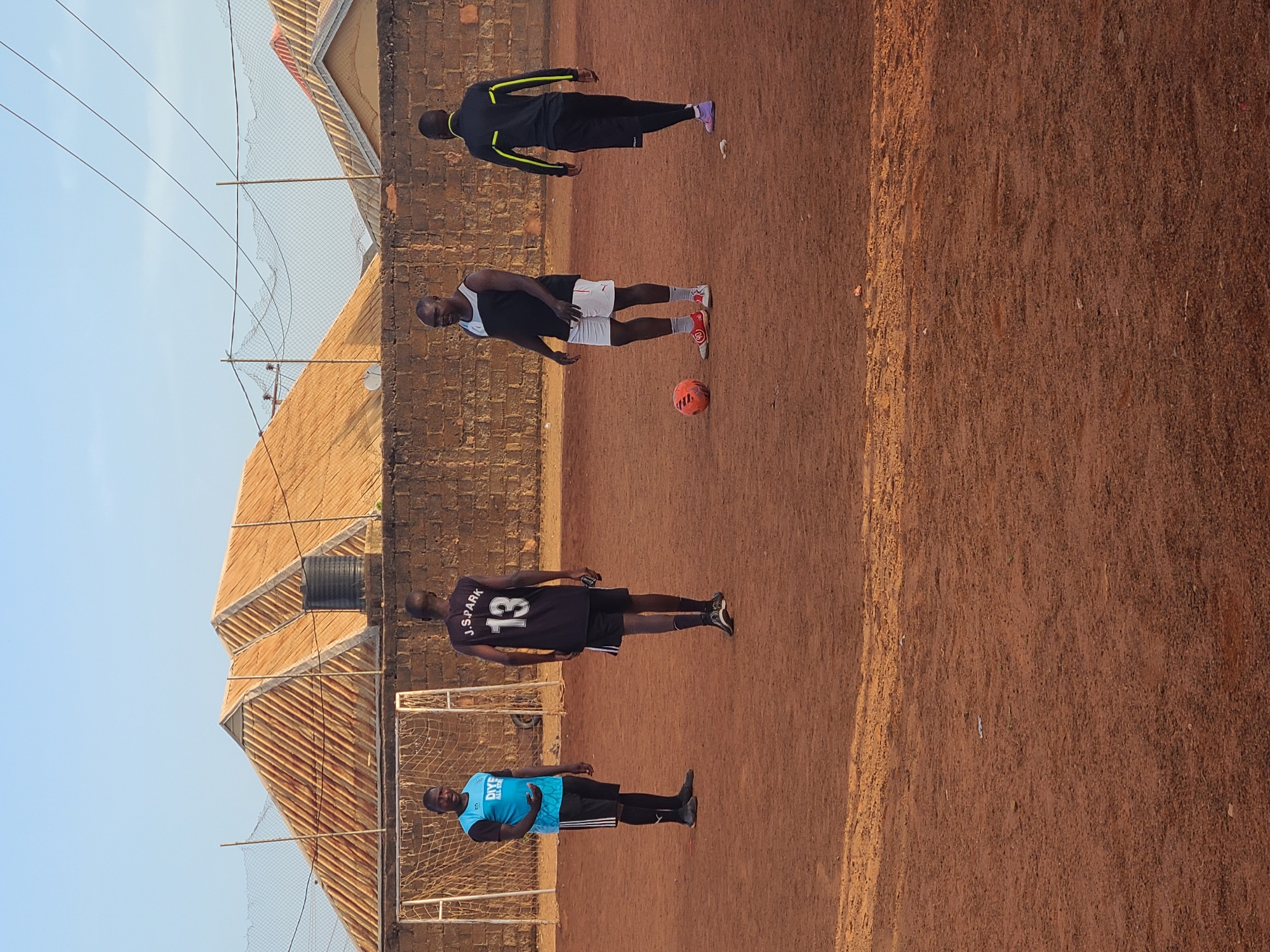 Four men are on a dirt soccer field. One man is wearing a black shirt and white shorts, and an orange soccer ball is near his feet. Another man is wearing a black shirt and pants with yellow accents. Another man is wearing a black shirt with "13 J. SPARK" on the back and black shorts. The fourth man is wearing a blue shirt and black shorts. There are soccer goals at either end of the field. Behind one goal is a brick building with a corrugated roof. The location is Unknown, Unknown.