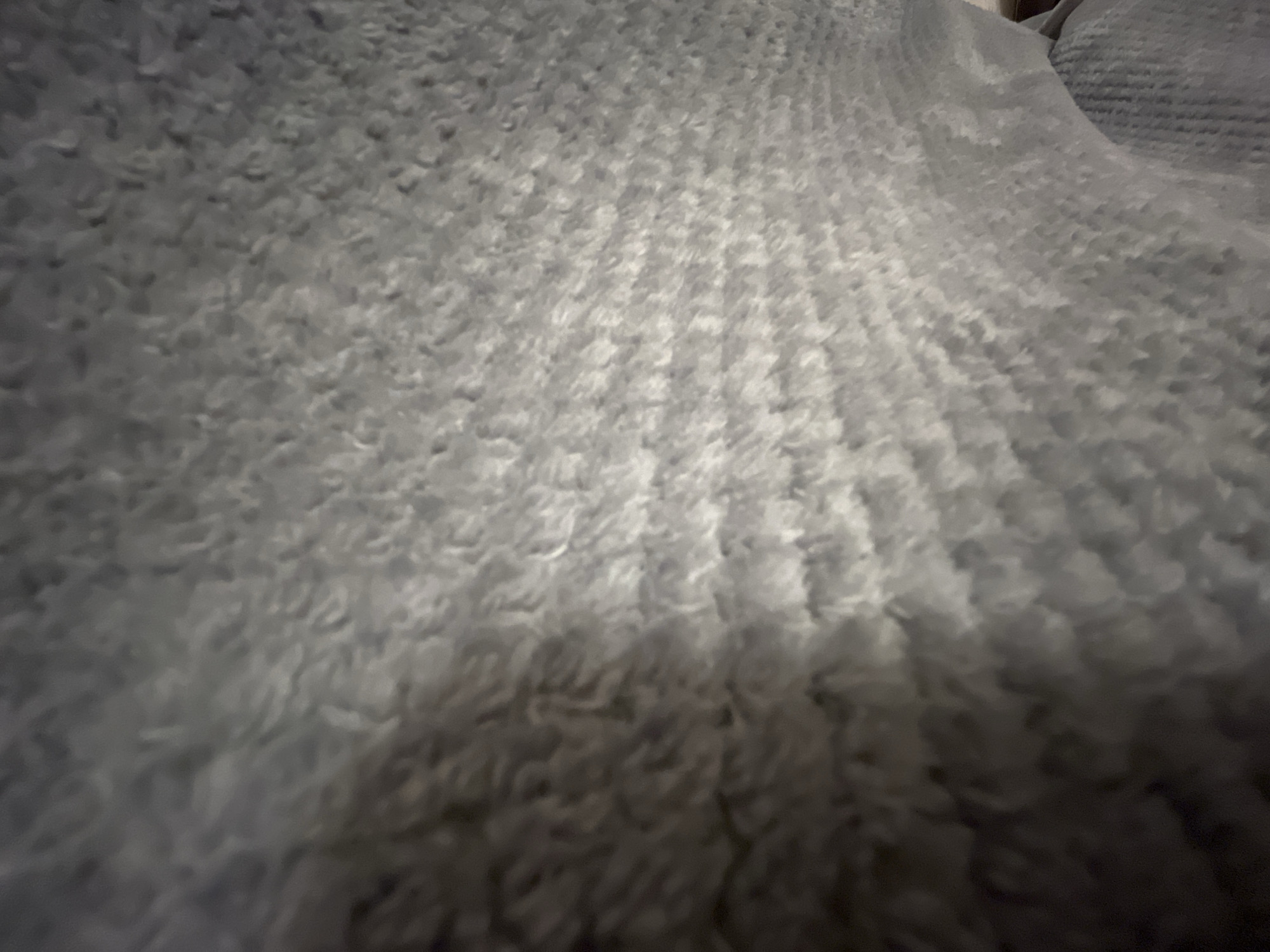The image is a close-up, low-angle shot of a soft, textured grey blanket. The blanket exhibits a ribbed pattern, suggesting it is knitted or woven. The lighting creates shadows and highlights, emphasizing the plush texture. The scene appears to be indoors, with no discernible background elements suggesting a specific location within Moscow, Russia. There are no people, objects, or actions depicted in the image.