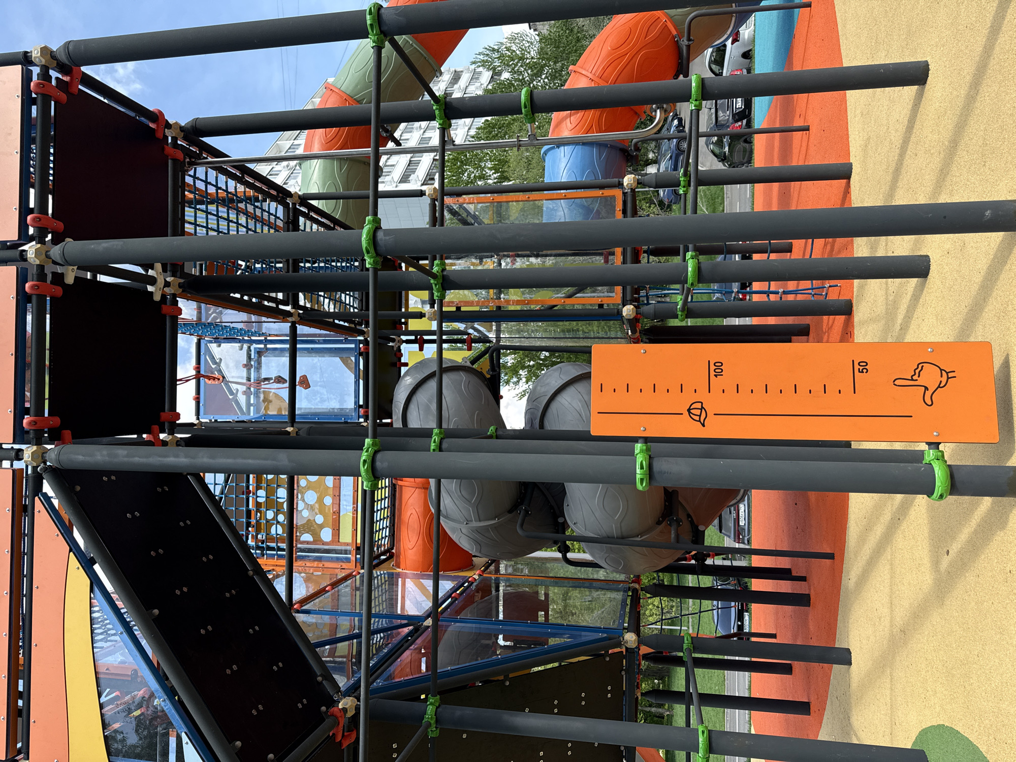 The image shows a brightly colored playground structure with interconnecting tubes, platforms, and climbing elements. The structure features gray metal pipes connected by green and orange joints. There are orange and green enclosed tubes, and sections with climbing nets. An orange sign marked with measurements, including the number 50, is attached to the structure. The ground surrounding the structure is covered in a soft, colorful material. A multistory building is visible in the background. The image was taken in Moscow, Russia.
