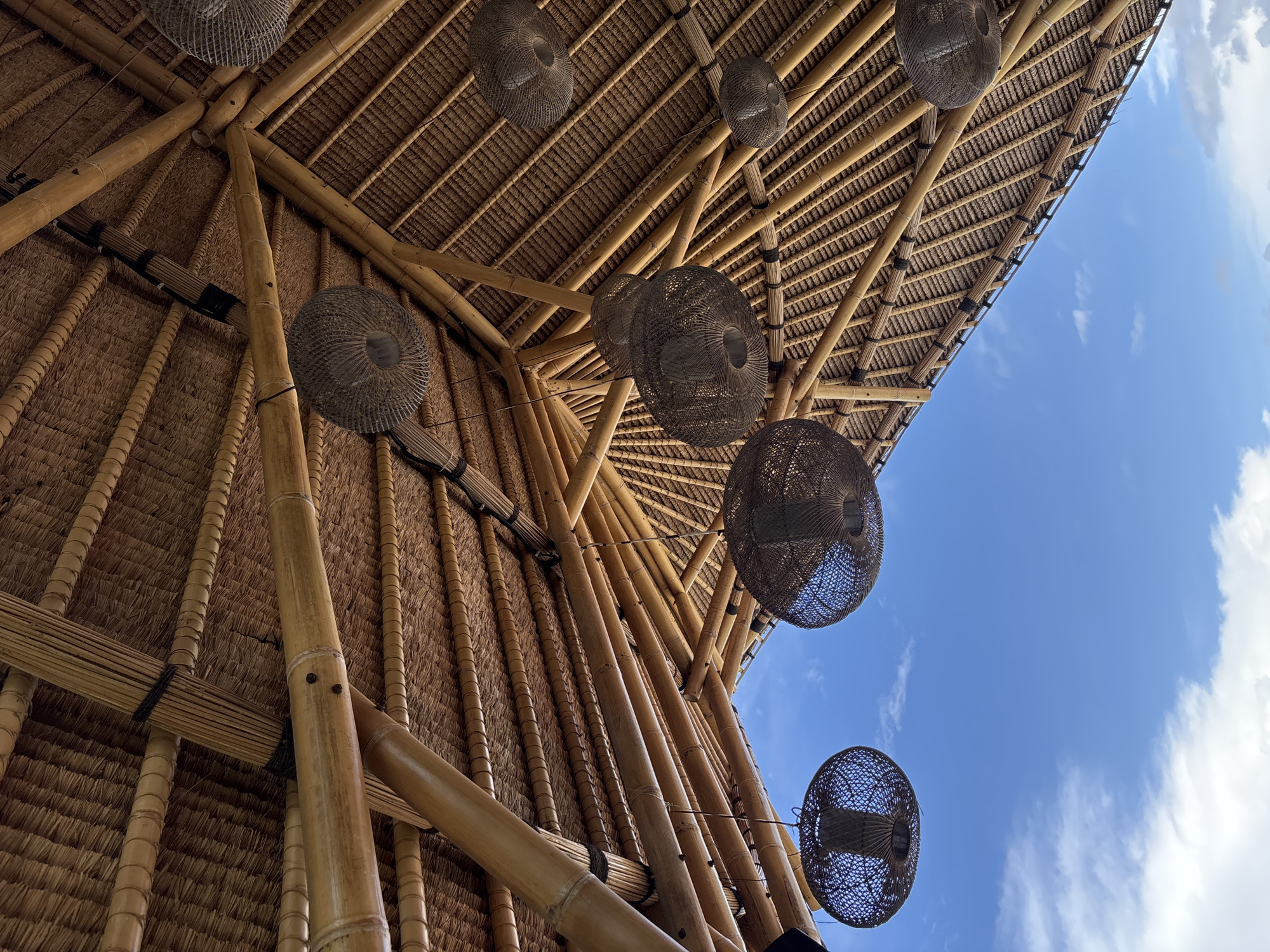 An upward-angle view captures the intricate ceiling structure of a building crafted from bamboo and natural thatch, set against a bright blue sky. The main structural elements consist of numerous robust, light-brown bamboo poles that serve as beams, rafters, and supports, forming a complex, intersecting network. Dark bindings are visible, securing the bamboo pieces together at various junctions.
The ceiling itself is a textured surface made from woven natural material, likely dried palm leaves or straw, arranged in neat, overlapping rows characteristic of traditional thatch roofing. Several dark, circular, woven pendant lights or lampshades, possibly made from rattan or wicker, hang down from the bamboo framework at different heights.
Through openings or from the edge of the roof structure, a clear blue sky with scattered white clouds is visible, suggesting an open-air design or a perspective looking outwards from beneath a canopy. The bright sky indicates it is daytime with good weather. The use of bamboo and thatch is typical of tropical architecture, fitting the location context of Kuta Selatan, Indonesia. No people or visible text are present in the image. The scene highlights craftsmanship and a natural, airy environment.