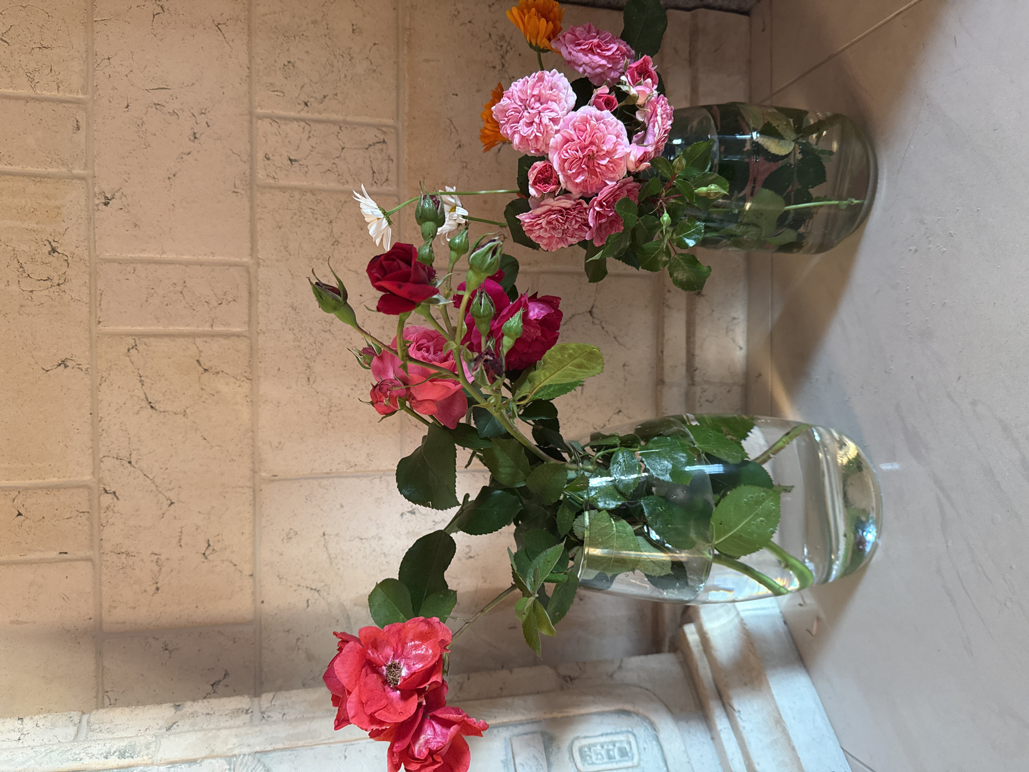 The image displays two glass vases filled with water and various flowers, positioned on a light-colored tiled surface. The vase on the left contains red roses and green leaves. The vase on the right holds pink roses, orange marigolds, and a few white daisies, also with green leaves. The background consists of a light-colored brick wall. To the left of the vases, a portion of a light-colored decorative column or architectural element is visible. The scene is indoors, under artificial or natural lighting that casts subtle shadows. There are no people or actions depicted in the image. The image does not provide any information about its location in the city of Saint Petersburg, Russia.