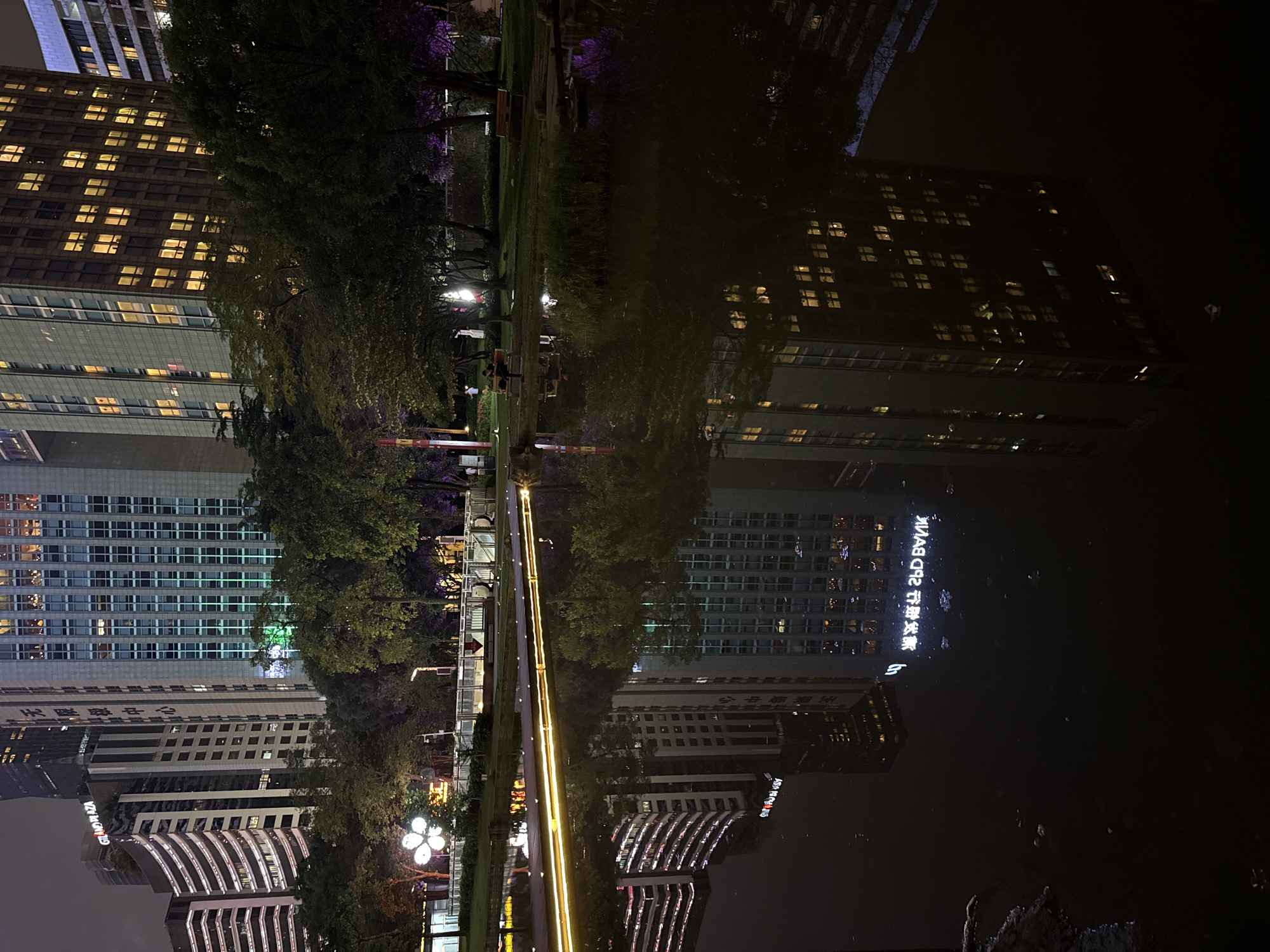The image shows a nighttime scene in Tianhe District, China, reflecting in a body of water. The reflection prominently features buildings with illuminated windows, with one building displaying the reversed text "26D BANK", as well as other reversed text in an Asian language. The illuminated cityscape is also reflected in the water. A park runs alongside the water, featuring trees and a string of lights that appear as a long, bright line in the image. People are barely visible in the park, possibly sitting on benches. The light is dim, and the dark water dominates the scene.