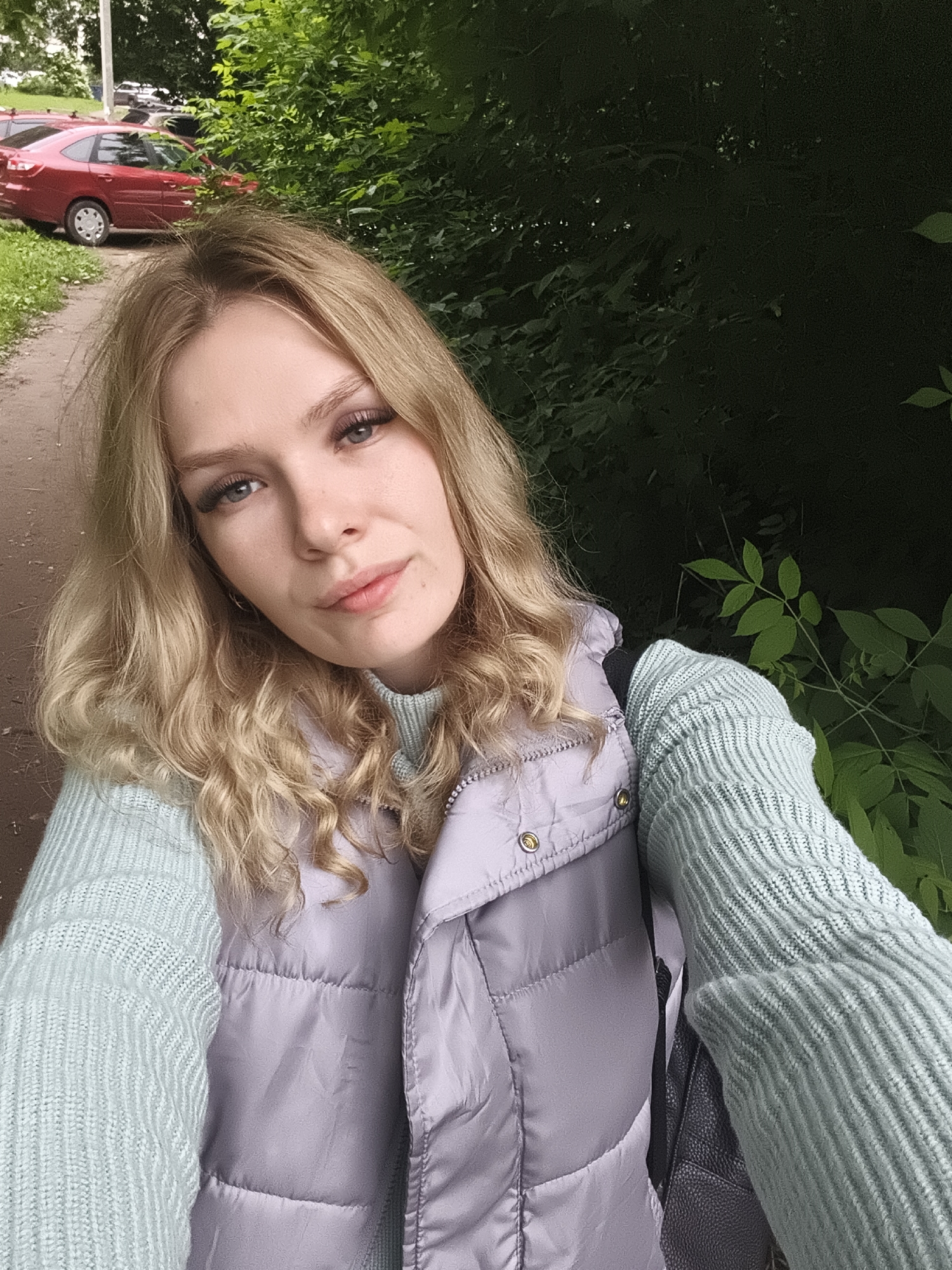 A young woman with long, wavy blonde hair and blue eyes is captured in a close-up selfie taken outdoors. She has a subtle, pleasant expression, looking directly into the camera. She is dressed in a light teal or mint green ribbed knit sweater and a light lavender quilted puffer vest. A black strap, likely from a backpack, is visible over her left shoulder.
The background reveals an outdoor setting, consistent with a public space in a city like Izhevsk, Russia. To the left, a dirt or gravel path is visible, alongside green grass and several parked cars, most notably a red sedan. To the right, dense green foliage from trees and bushes creates a natural backdrop. The lighting suggests it is daytime, possibly overcast or late afternoon, with soft, diffused light. There is no visible text in the image.