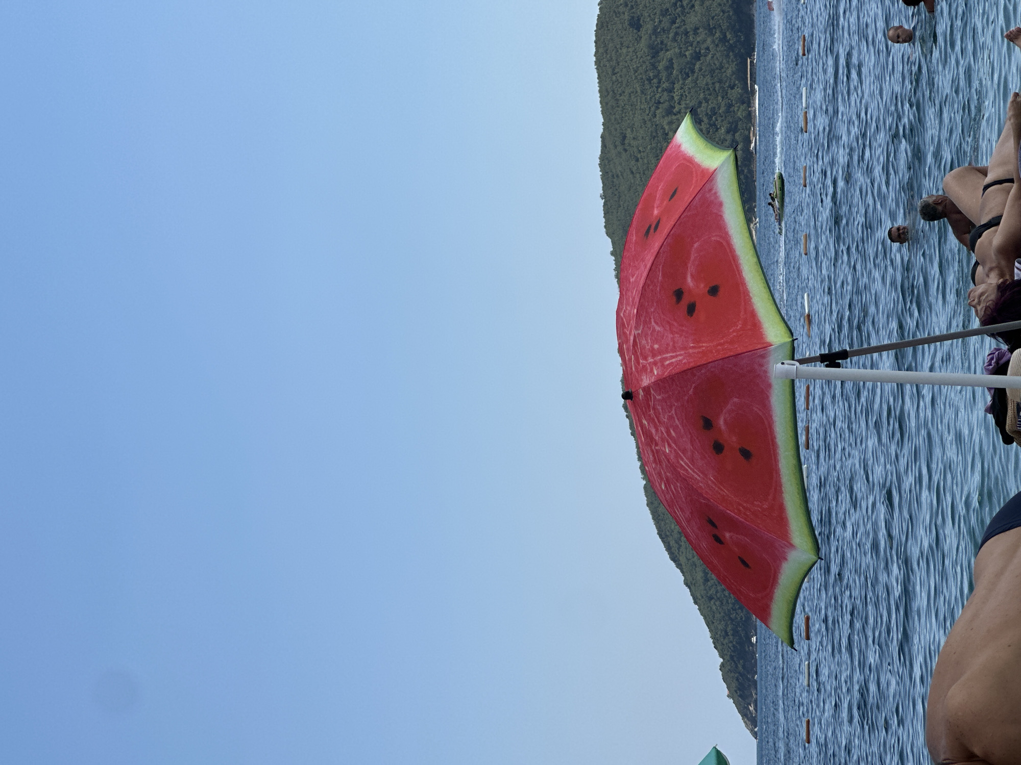 The image, taken in Budva, Montenegro, depicts a beach scene with people enjoying the water. A vibrant, watermelon-themed umbrella is the central object, open and casting shade. The umbrella's design features a bright red interior with black seeds and a green rind. In the background, a lush green hillside rises from the sea, suggesting a coastal mountainous landscape. The water is a deep blue, rippled by a gentle breeze, and several buoys are visible floating on the surface, marking a designated swimming area.
Several individuals are partially visible in the water and on the shore. Some are submerged to their necks, indicating they are swimming, while others appear to be lounging or preparing to enter the water. The overall atmosphere suggests a warm, sunny day at the beach, likely during daylight hours. The clarity of the sky and the activity of the people imply pleasant weather. The focus is on leisure and recreation in a beautiful natural setting. No text is visible in the image.