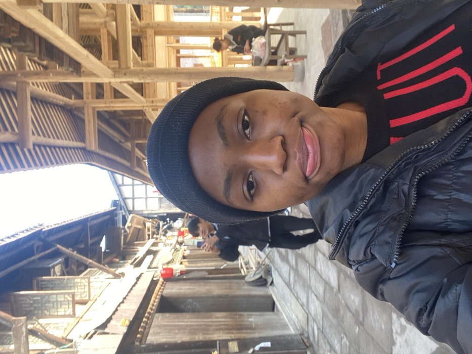 A selfie captures a person wearing a black beanie, a black sweater with red stripes, and a black jacket. They're standing outside, possibly in a market or village. Traditional wooden architecture is visible in the background, including roofs and walkways. Other people can be seen walking or sitting in the background, some appearing to be engaged in daily activities. The lighting suggests it is daytime.
