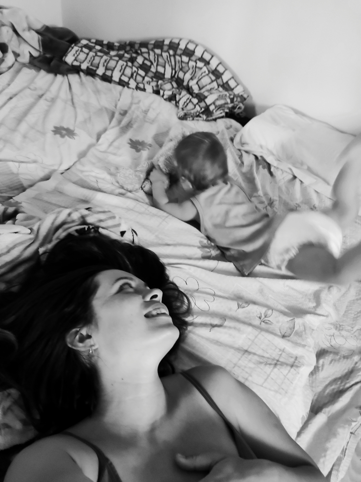 A monochrome shot shows a woman lying on her back on a bed, looking upward and smiling, with a baby crawling towards her. The woman has dark hair and is wearing a tank top. The baby, in a light-colored onesie, is positioned near a pillow. In the background, the bed has patterned sheets and a folded blanket. The location is an indoor bedroom.