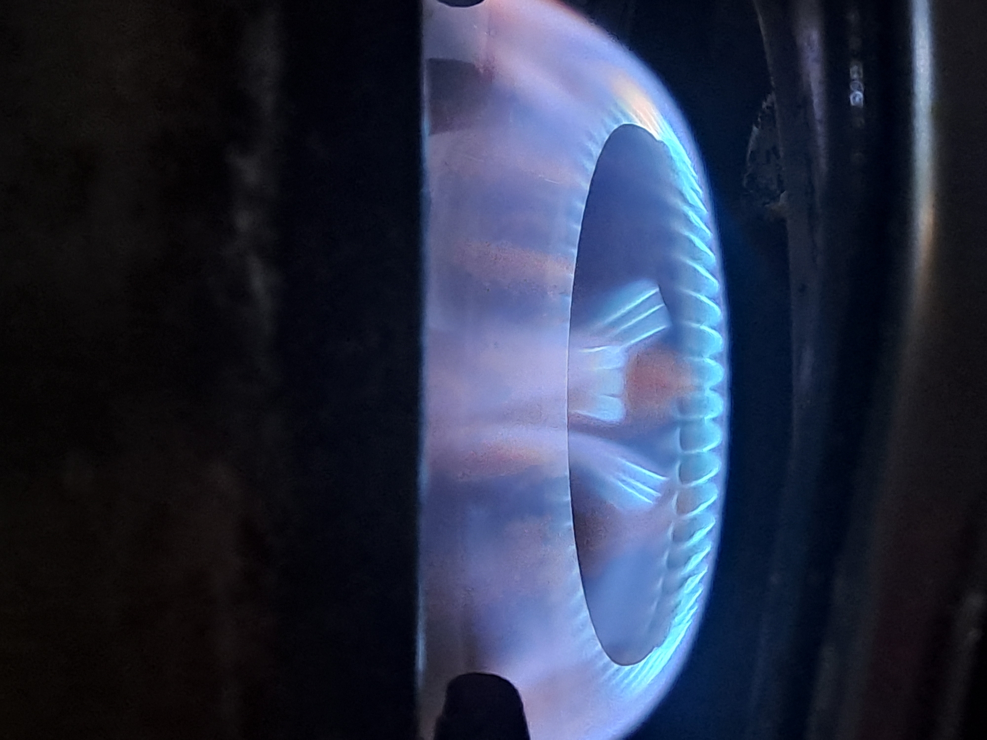The image shows a close-up view of a gas burner on a stove, with a bright blue flame. The flame is the primary subject, emitting a soft glow that illuminates the surrounding grate. The burner is circular, with a series of small holes from which the gas is escaping and igniting. The background is dark, suggesting the image was taken in low light conditions. There are no people or other objects visible in the scene. The location in the city of Sumberejo, Indonesia is not discernible from the image itself.