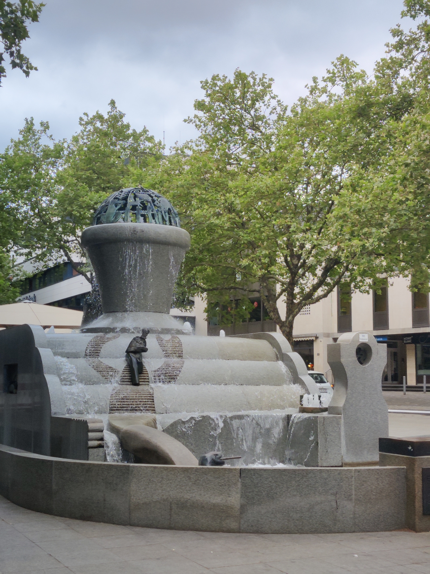The image shows the "Winckelmann Fountain" in Dortmund, Germany, located in front of the Reinoldikirche. The fountain is made of concrete and stone, with water flowing down multiple tiers. At the top of the fountain is a large, bowl-shaped structure topped with a metal sphere, resembling a globe, adorned with various figures. Below this, the fountain cascades down in a series of steps and basins. In the foreground, a bronze sculpture of a man is depicted climbing the fountain. The surrounding environment includes mature trees with green foliage, buildings with large windows, and a paved ground area. The sky is overcast.