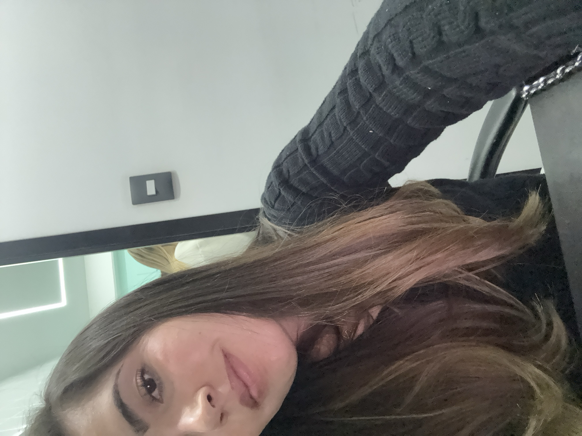 The image depicts an indoor scene in San Bernardino Tlaxcalancingo, Mexico, centered on a young adult female captured in a close-up, front-facing perspective. She has long, straight brown hair with lighter, possibly ombré, ends, falling over her shoulders and chest. Her skin tone is light, and she has visible eyebrows, dark eyes, and a slight, closed-mouth smile. She is wearing a dark, likely black, long-sleeved top with a distinct ribbed or cable-knit texture.
In the background, to the subject's right, a clean white wall is visible, featuring a grey light switch with a white rectangular toggle. Above and to the left of the subject's head, a modern linear white light fixture is visible on the ceiling. A reflection of another individual with blonde hair wearing a white garment is partially visible in a reflective surface or mirror behind the primary subject. The lower right portion of the image shows part of a dark, possibly office-style, chair and a dark object with a silver chain strap, consistent with a purse or bag, suggesting the subject is seated at a desk or table. The environment appears to be a modern, minimalist interior.